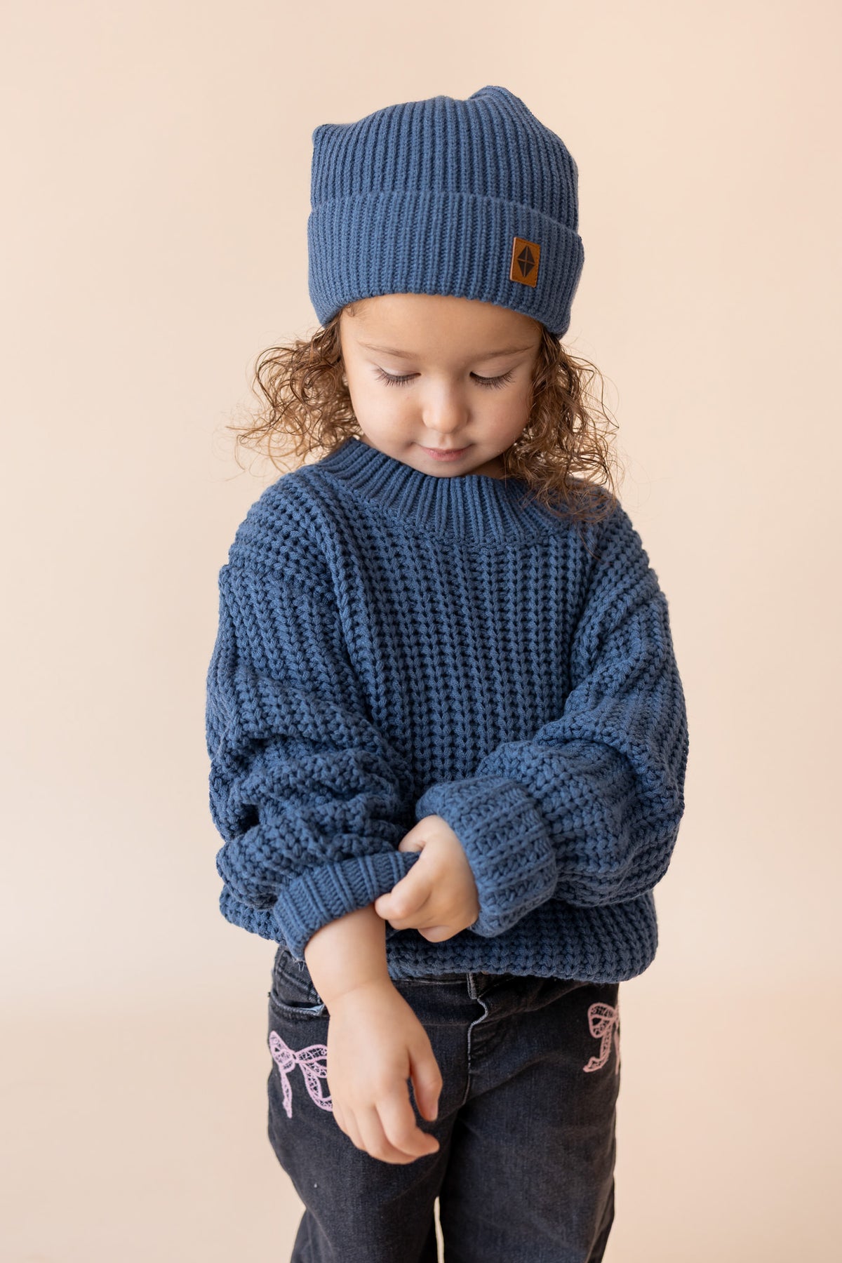 Girl wearing Chunky Knit Sweater and chunky knit pom beanie in Oxford over black pants on a beige background