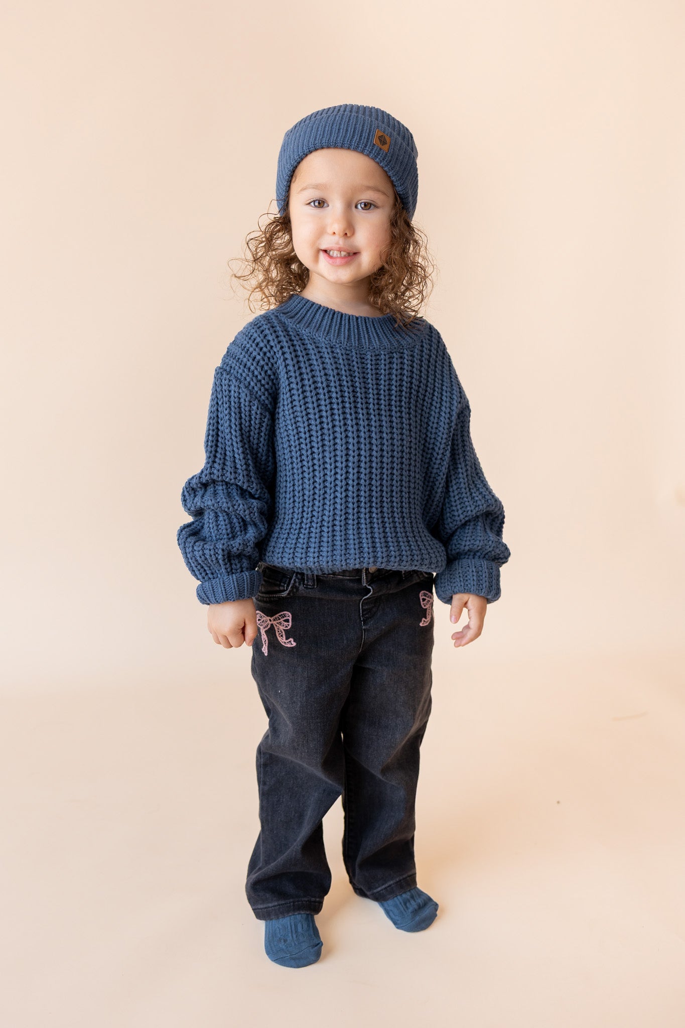 Child wearing chunky knit sweater and chunky knit pom beanie in oxford over black pants on a beige background