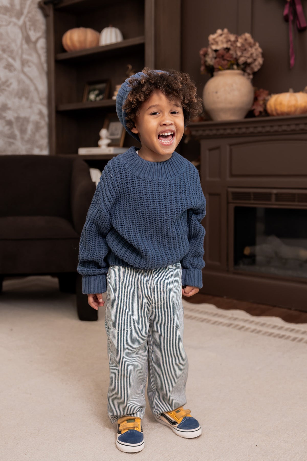 Bow wearing Chunky Knit Sweater and chunky knit pom beanie in Oxford over white pants standing in a living room
