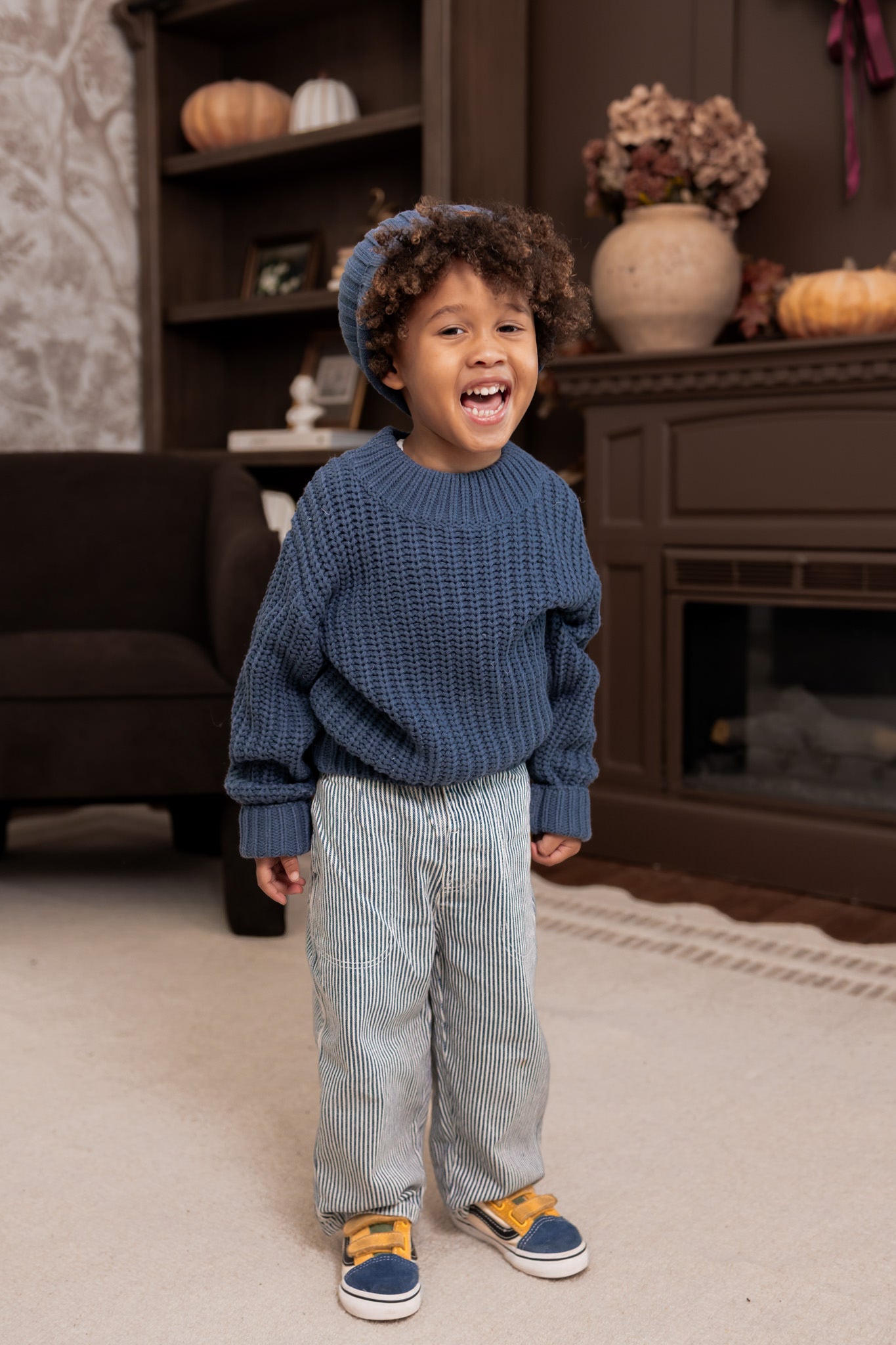 A young child wearing a chunky blue knit sweater, light blue striped pants, and blue sneakers with yellow accents, standing in a cozy living room setting.