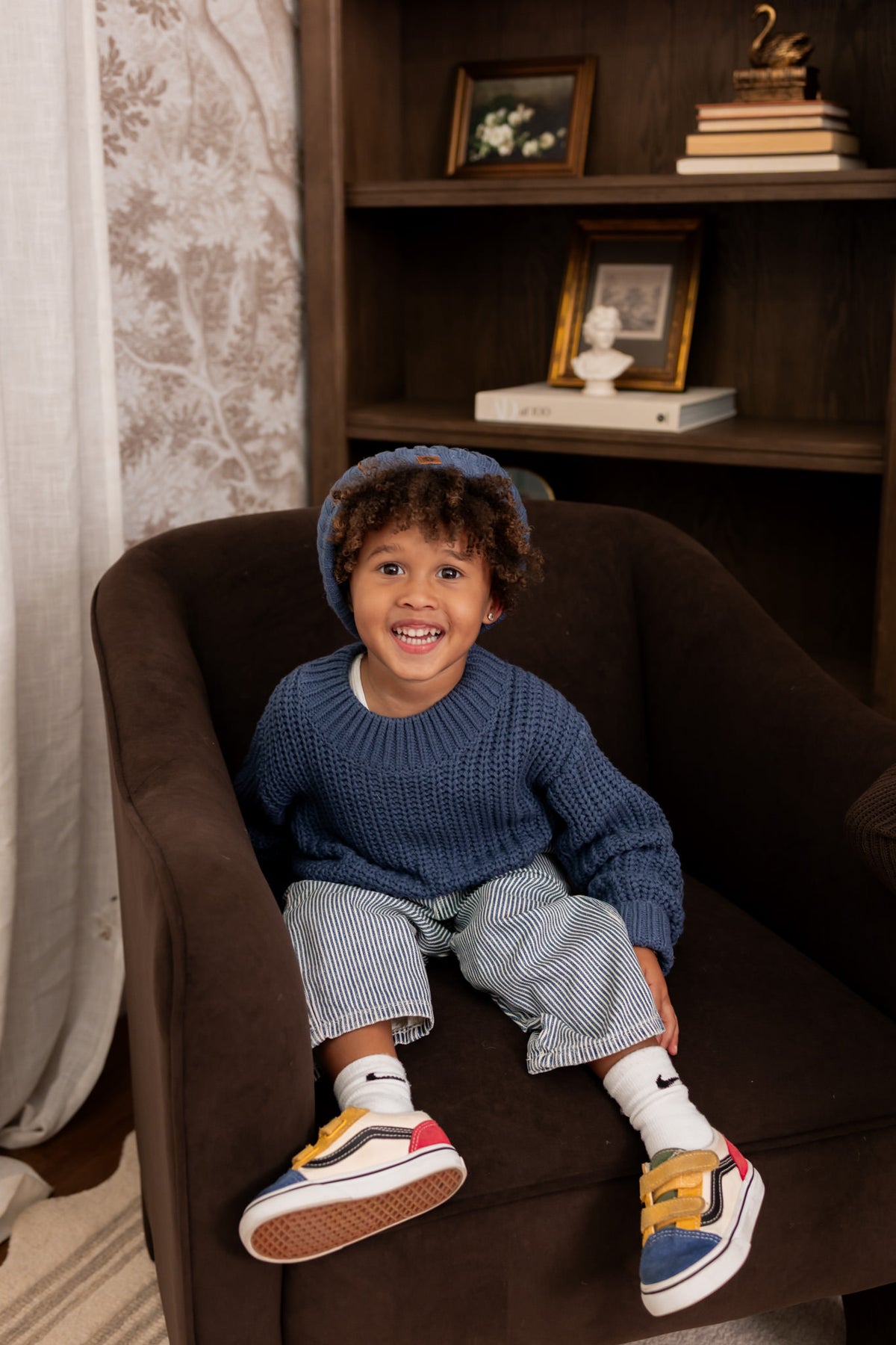 Boy sitting on brown sofa wearing Chunky Knit Sweater and chunky knit pom beanie in Oxford over white pants