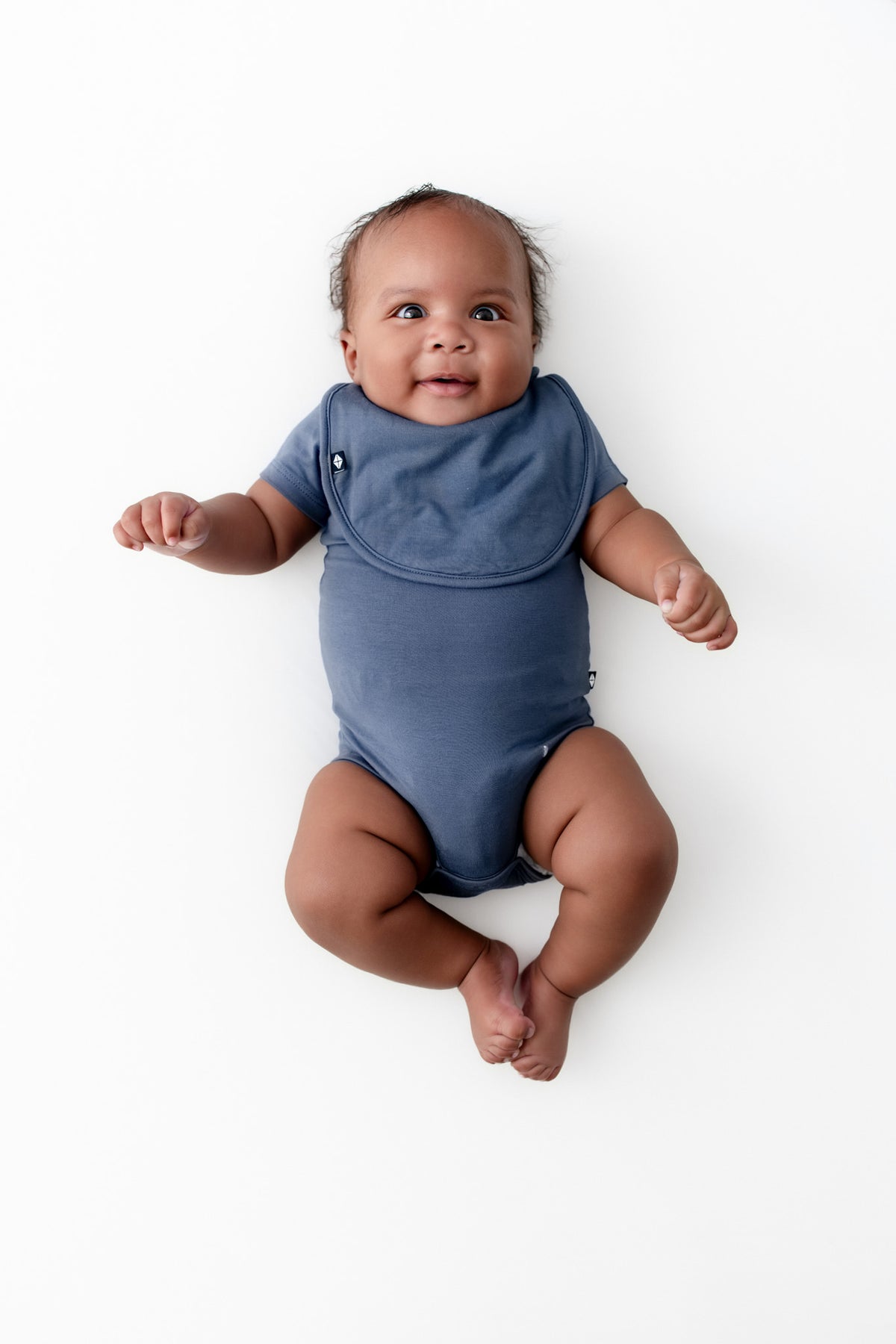 Baby wearing Cushy Bib in Oxford over similar color bodysuit on a white background