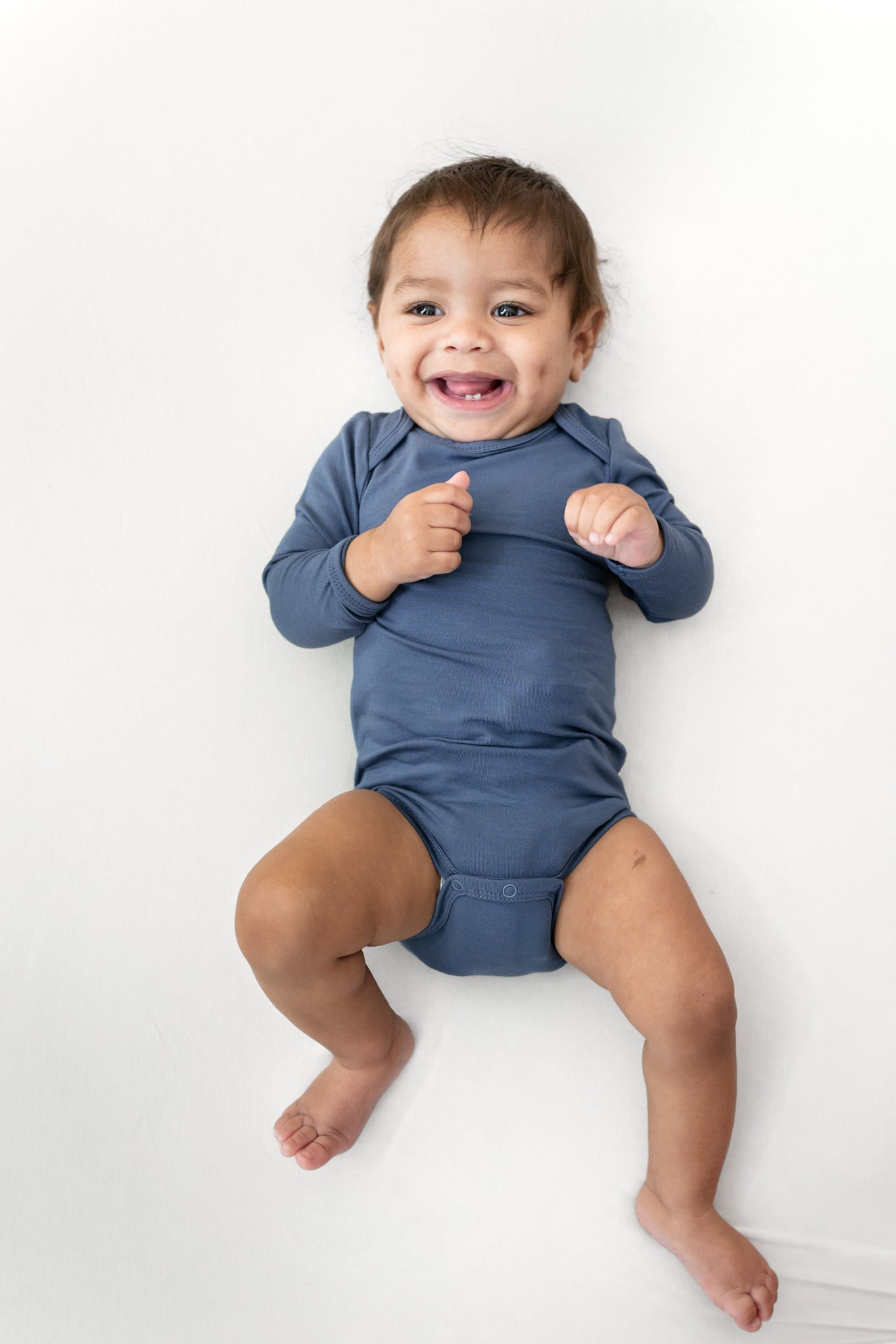 A smiling baby lying on a white background, wearing a long-sleeve blue bodysuit.