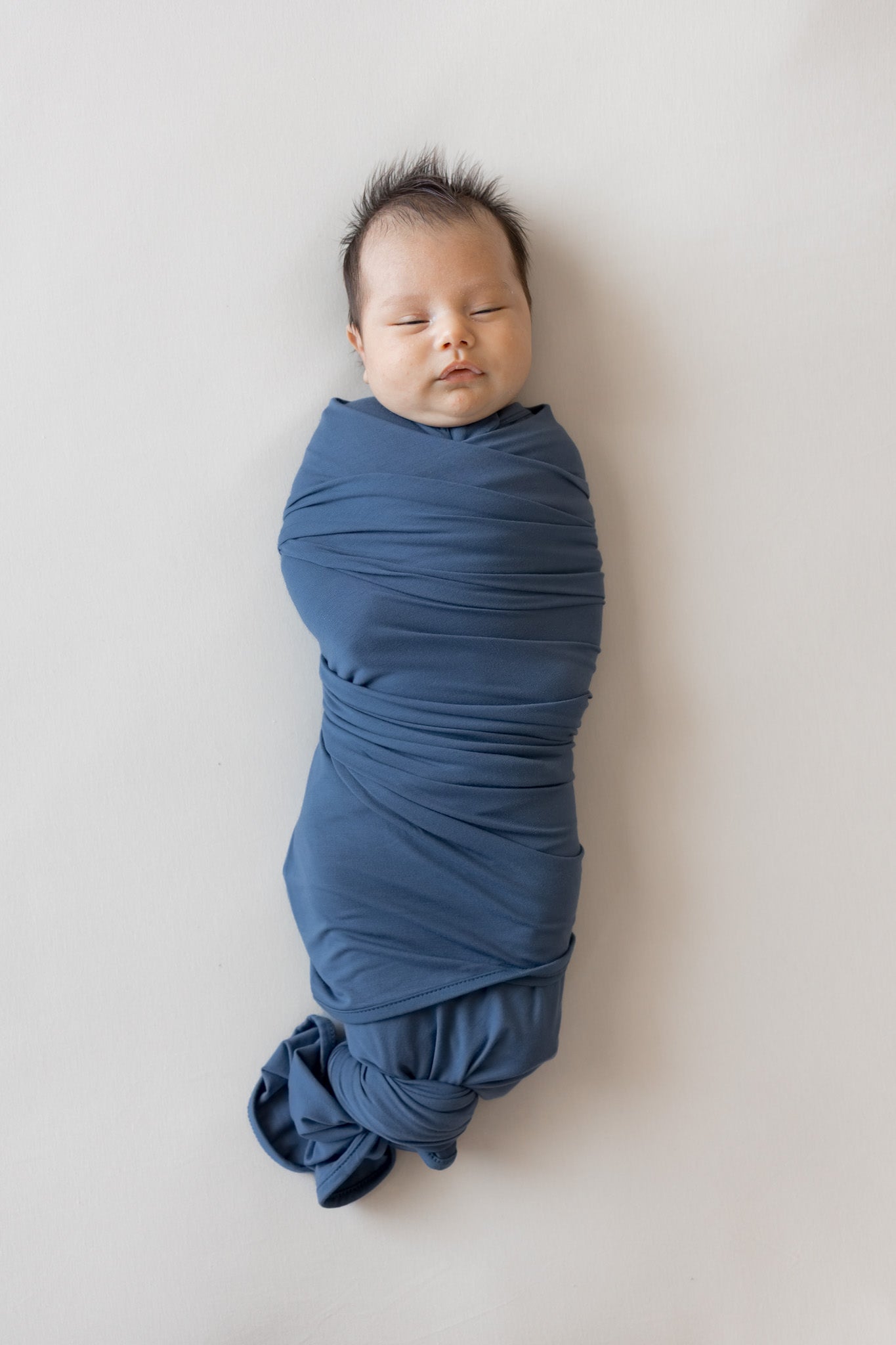 A sleeping newborn baby swaddled in a dusty blue fabric wrap, lying on a light beige background.