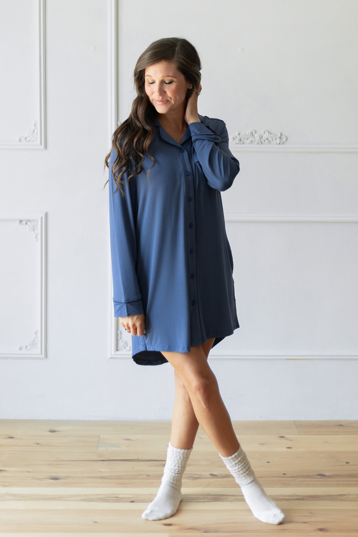 A woman wearing a slate blue button-down shirt dress and white socks, standing on a light wood floor against a white paneled wall.