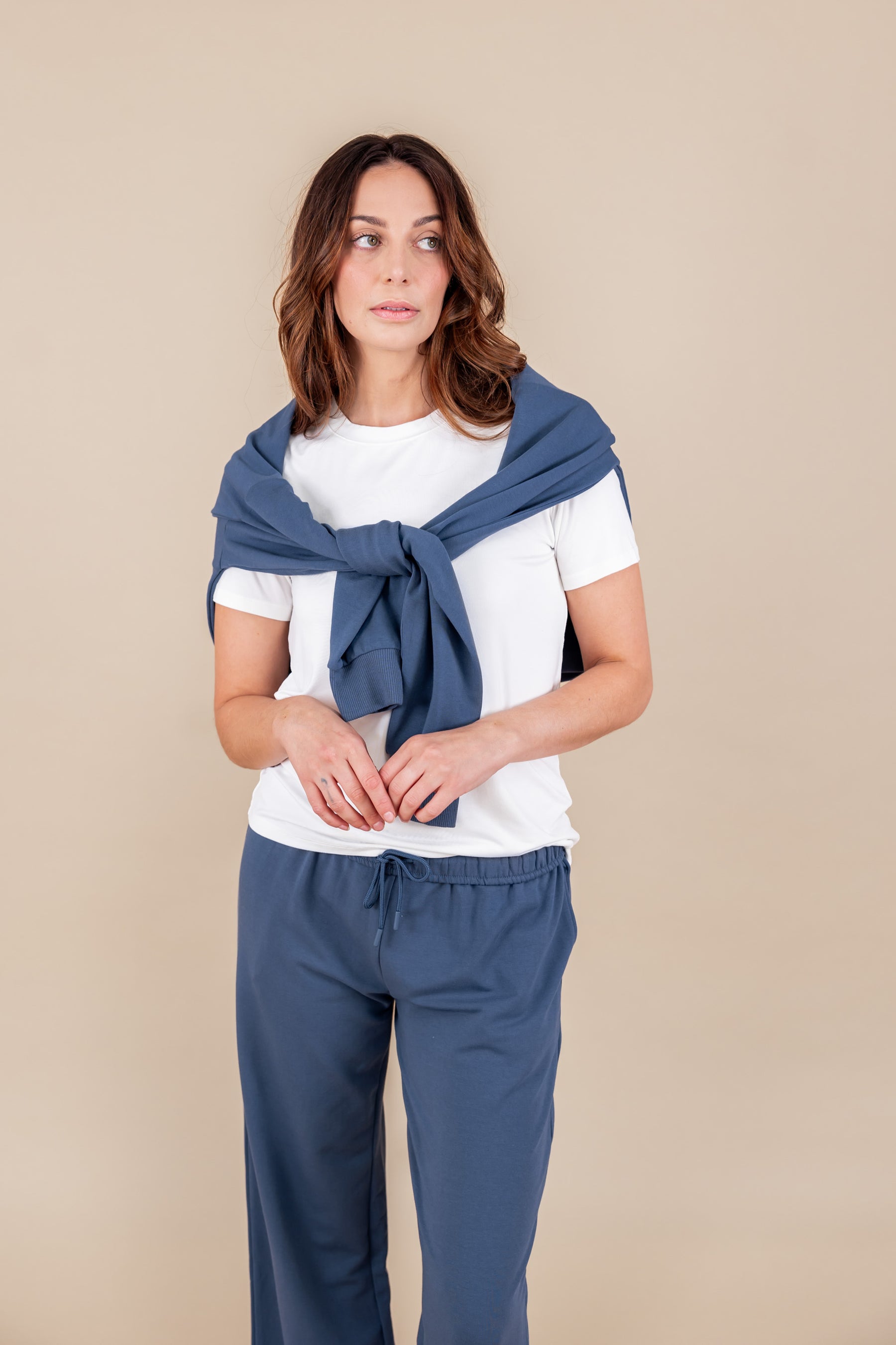 Woman wearing a white T-shirt and Women's Hoodie in Oxford color over her shoulders over similar color sweatpants on a beige background