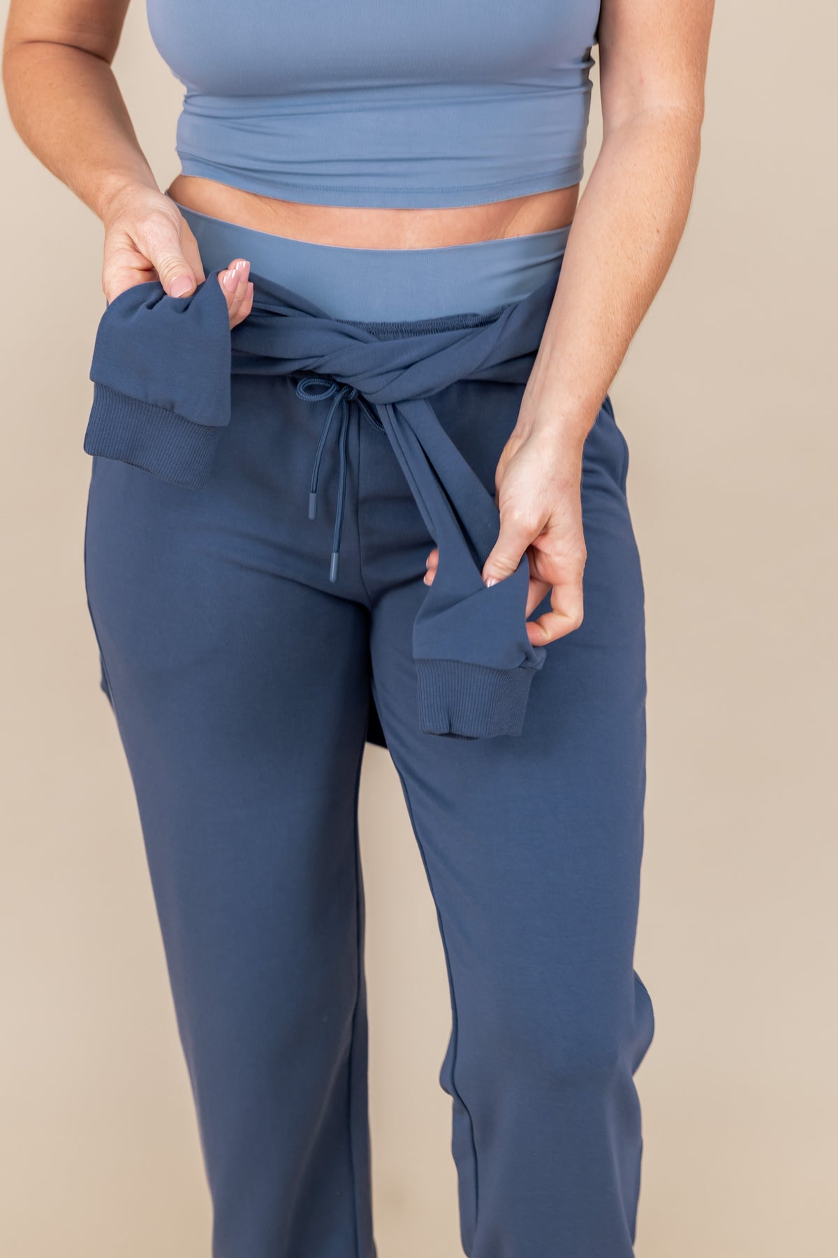 Modal wearing Women's Jogger Pants in Oxford color under flex Bra tank in breeze while tying a oxford color sweatshirt on her waist with the sleeves on a beige background