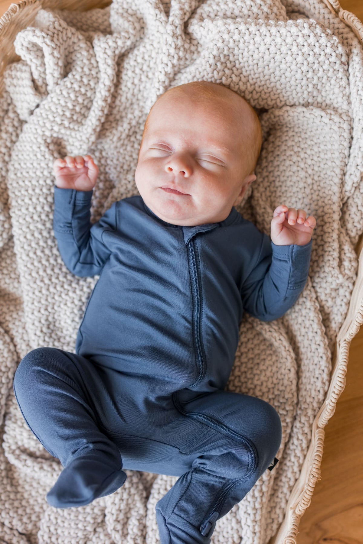 A sleeping newborn baby wearing a navy blue footed zip-up onesie, lying on a beige knit blanket inside a wicker basket.