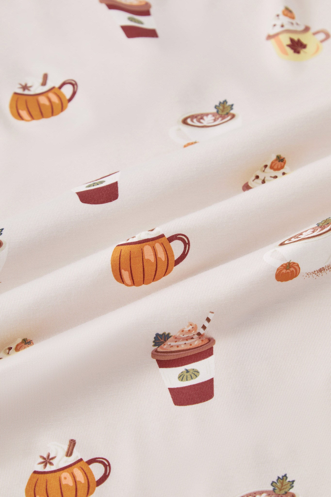 A repeating, all-over print featuring: a white cappuccino cup with latte art and a green leaf garnish, next to a mini pumpkin and a dusting of cocoa powder; an orange pumpkin-shaped mug with whipped cream and garnished with star anise and a cinnamon stick; a rust-colored to-go coffee cup with orange whipped cream, a dark green leaf, and a straw; and a yellow mug with a maple leaf image, topped with whipped cream, cloves, and a mini orange pumpkin, over a light beige background color named Vanilla.