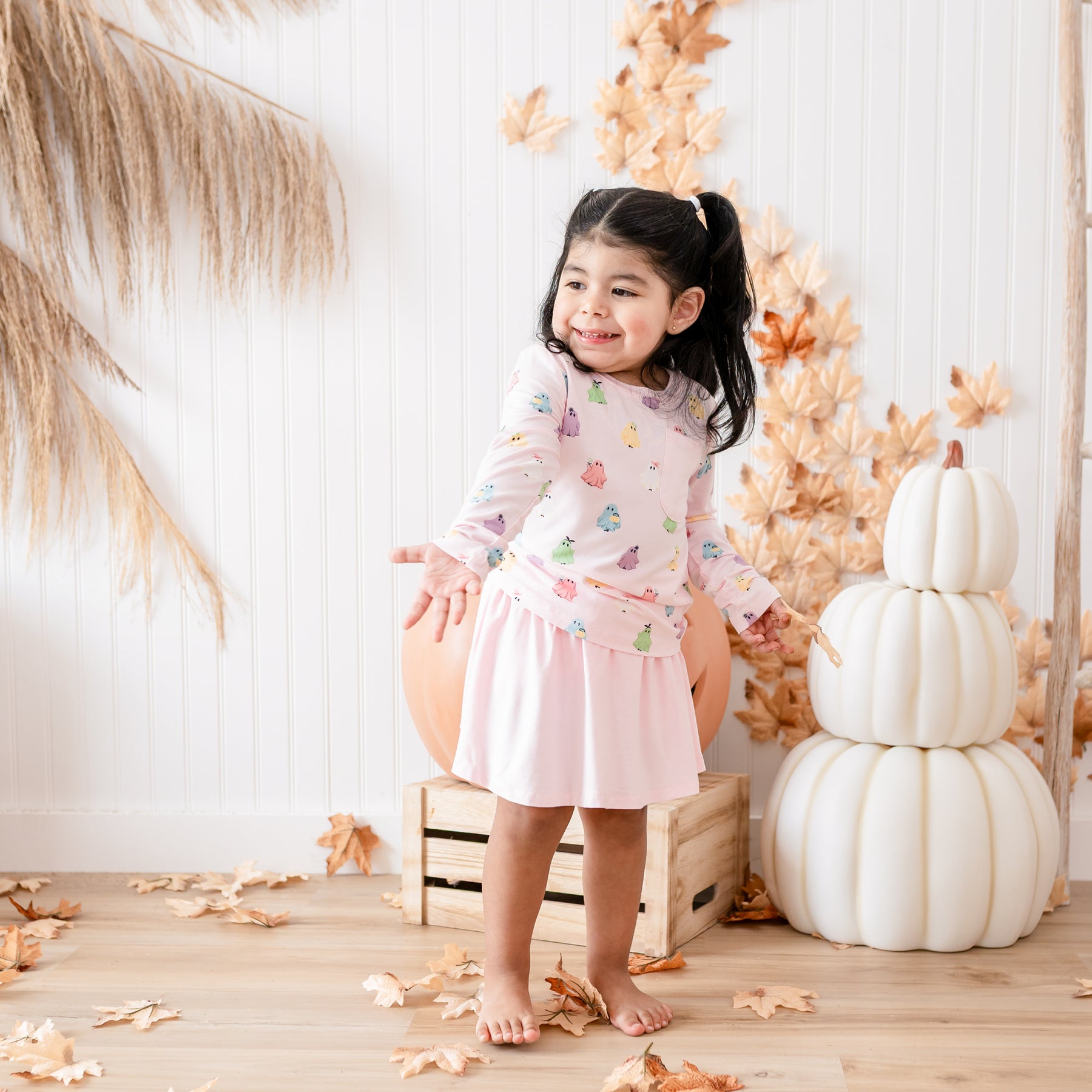 Toddler shrugging her shoulders wearing the Long Sleeve Toddler Crew Neck Tee in Pastel Ghost paired with a Sakura skort in front of white stacked pumpkins, leaves and a wooden crate