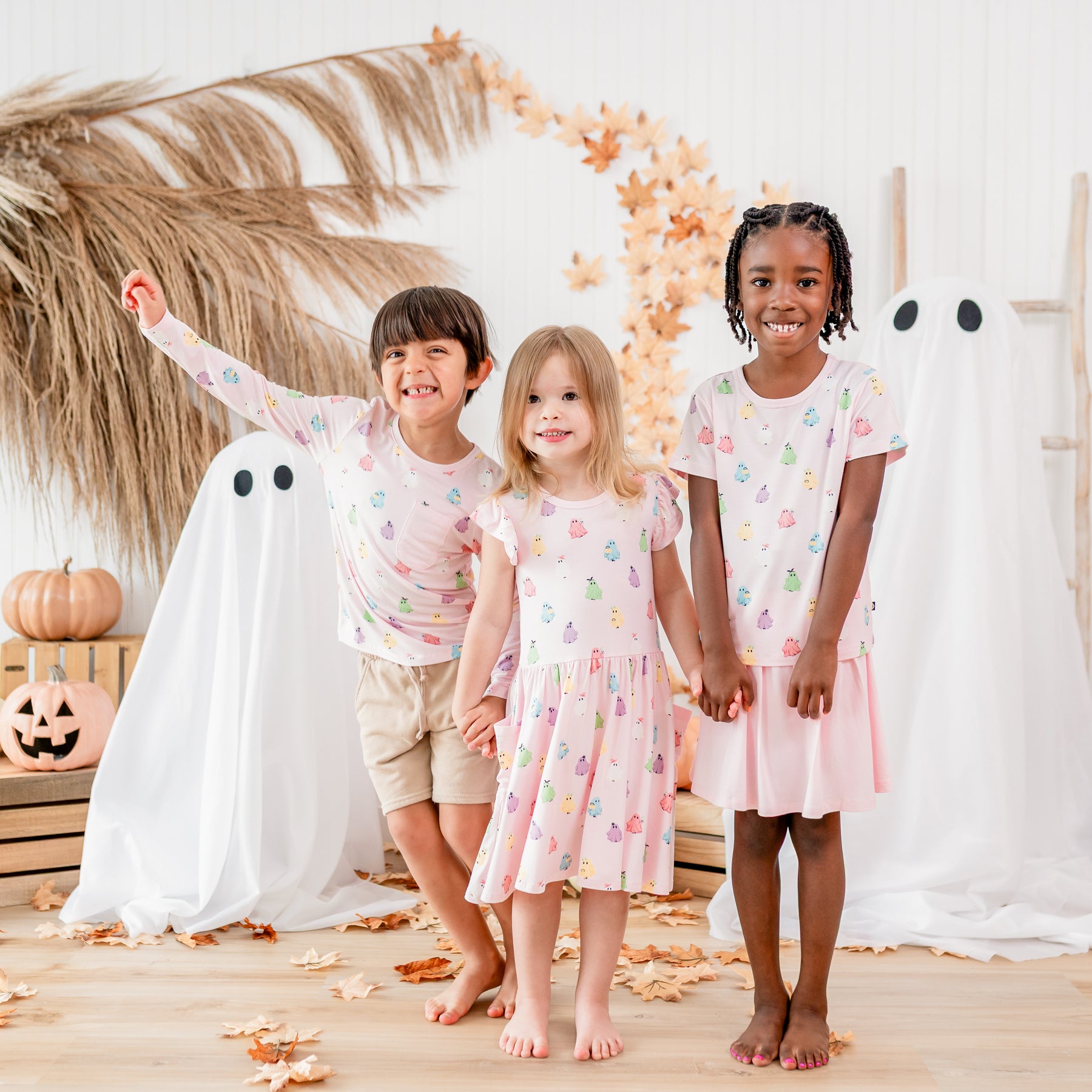 Three children standing holding hands matching in the Bewitched print from the Halloween collection in front of sheet ghosts, leaves and pumpkins