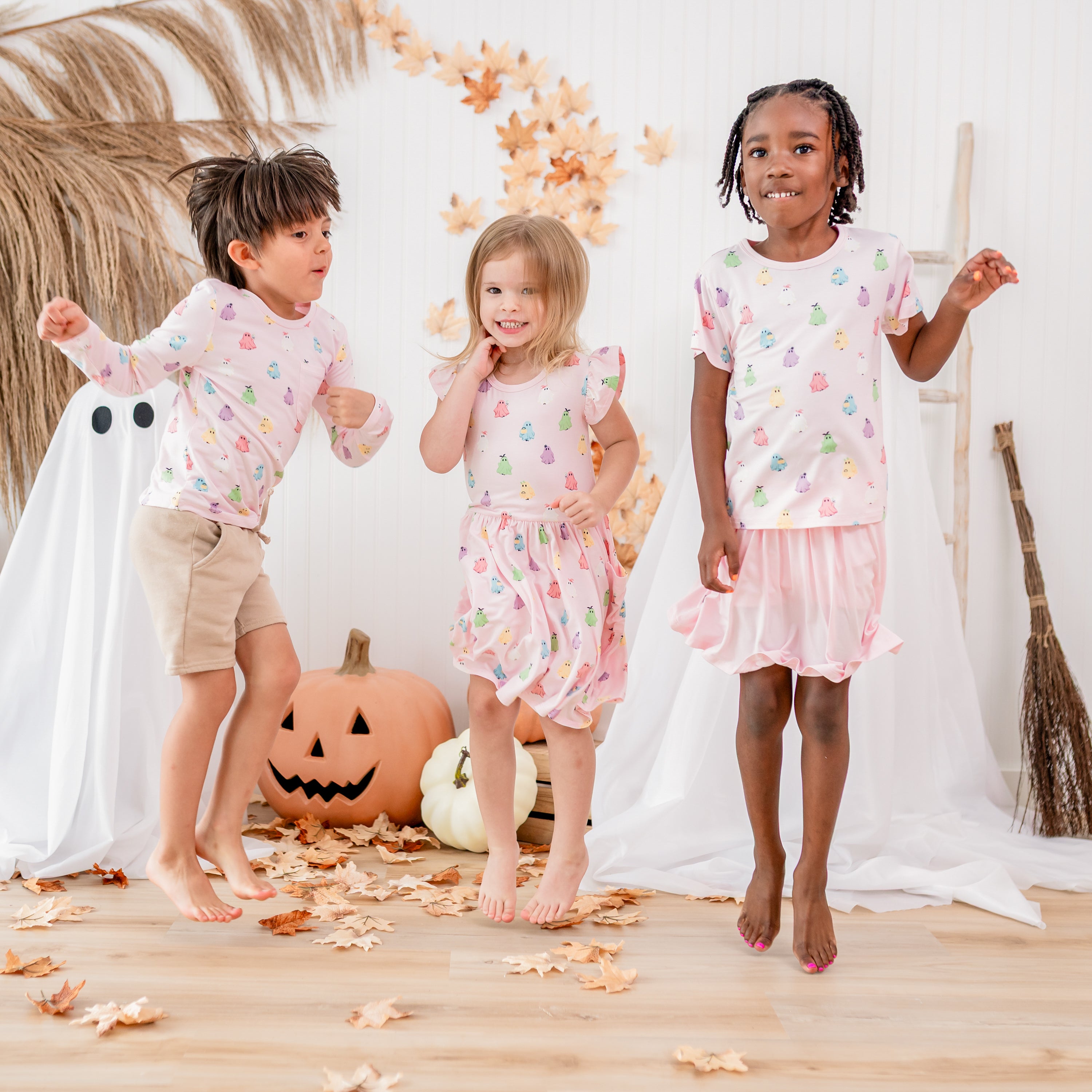 Three kids jumping in the air in front of a fall backdrop wearing various items in Pastel Ghost from the Halloween collection. 