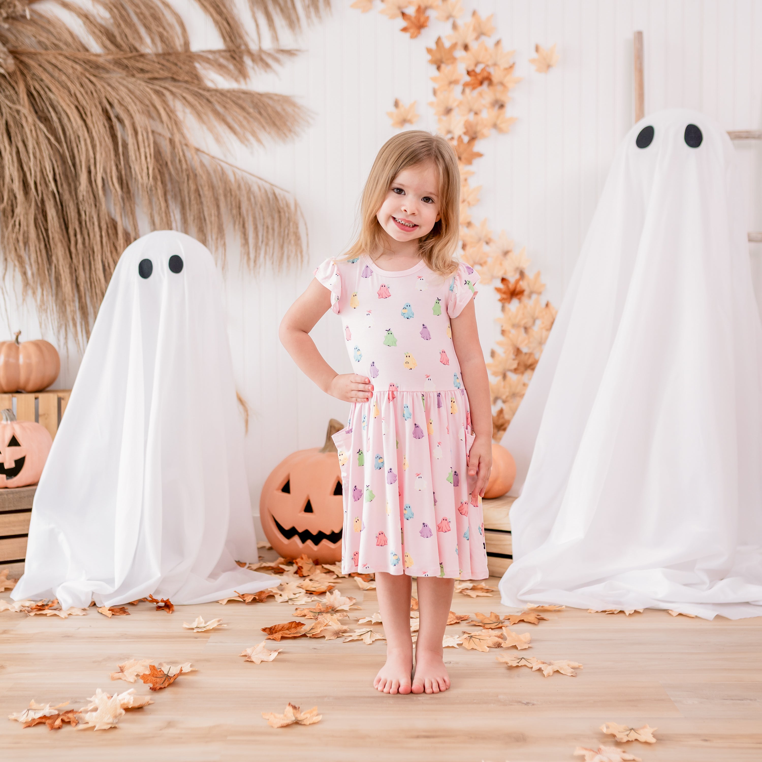 Young girl standing with one hand on her hip in front of a fall backdrop of sheet ghosts, leaves, pumpkins and wooden crates wearing the Pocket Dress in Pastel Ghost
