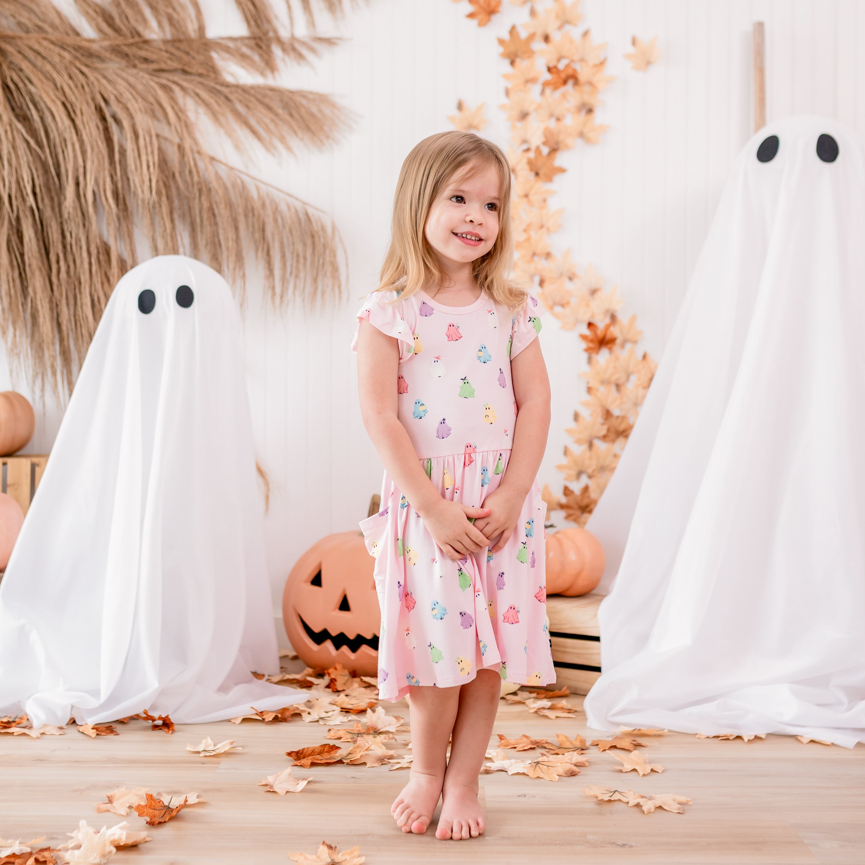 Young girl clasping her hands together wearing the Pocket Dress in Pastel Ghost in front of a fall inspired backdrop of sheet ghosts, pumpkins, leaves and wooden crates