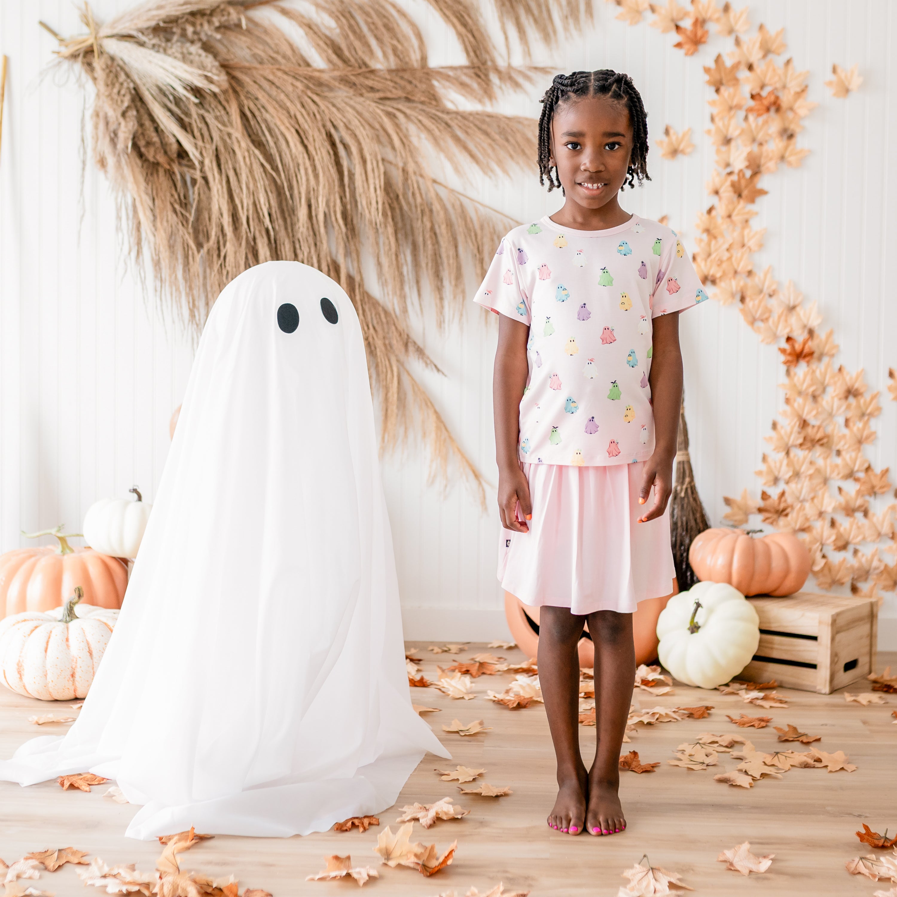 Smiling girl standing beside a sheet ghost in front of a fall backdrop wearing the Toddler Basic Tee in Pastel Ghost paired with a Sakura Skort