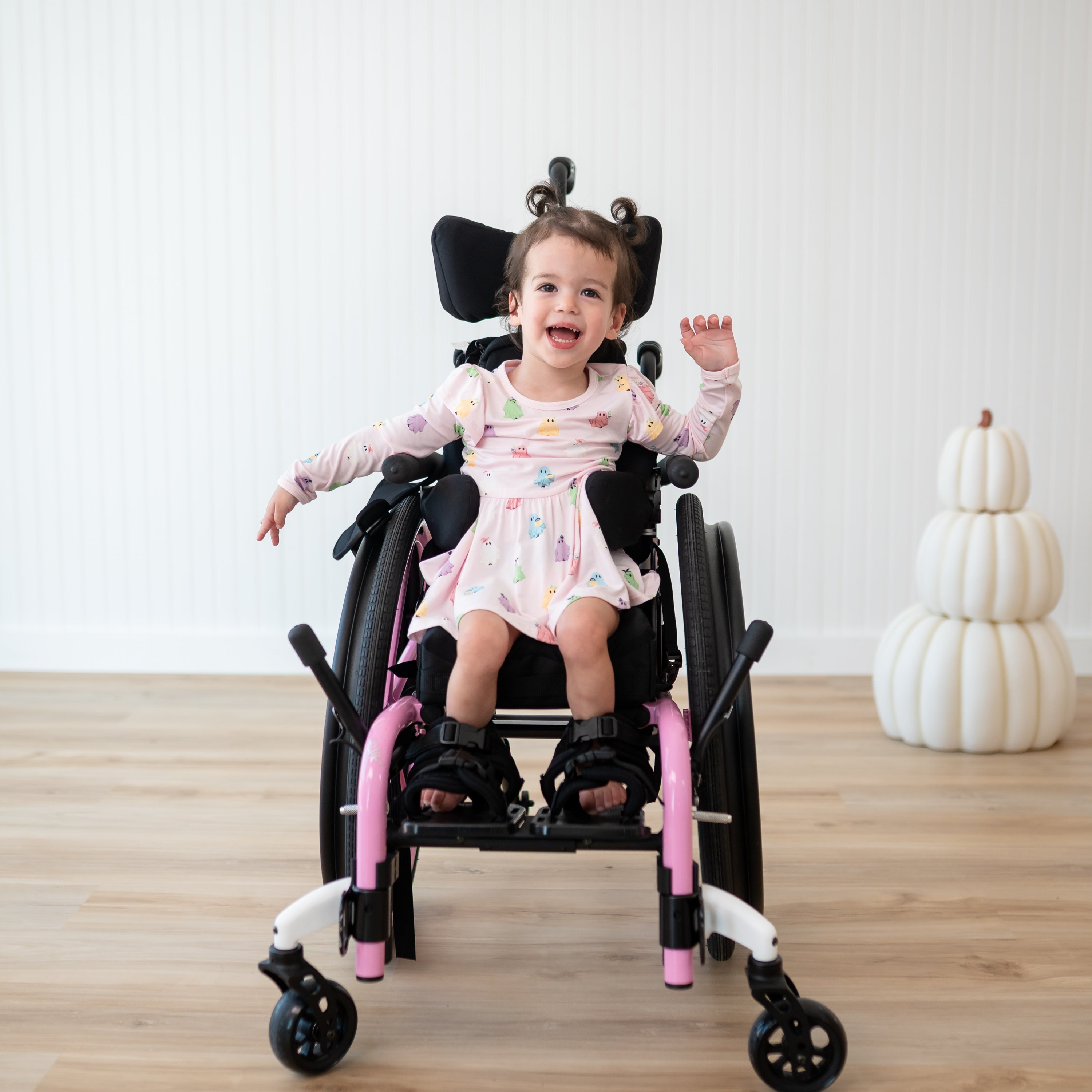 Young girl smiling sitting in her wheel chair beside a stack of white pumpkins wearing the Long Sleeve Twirl Bodysuit Dress in Pastel Ghost
