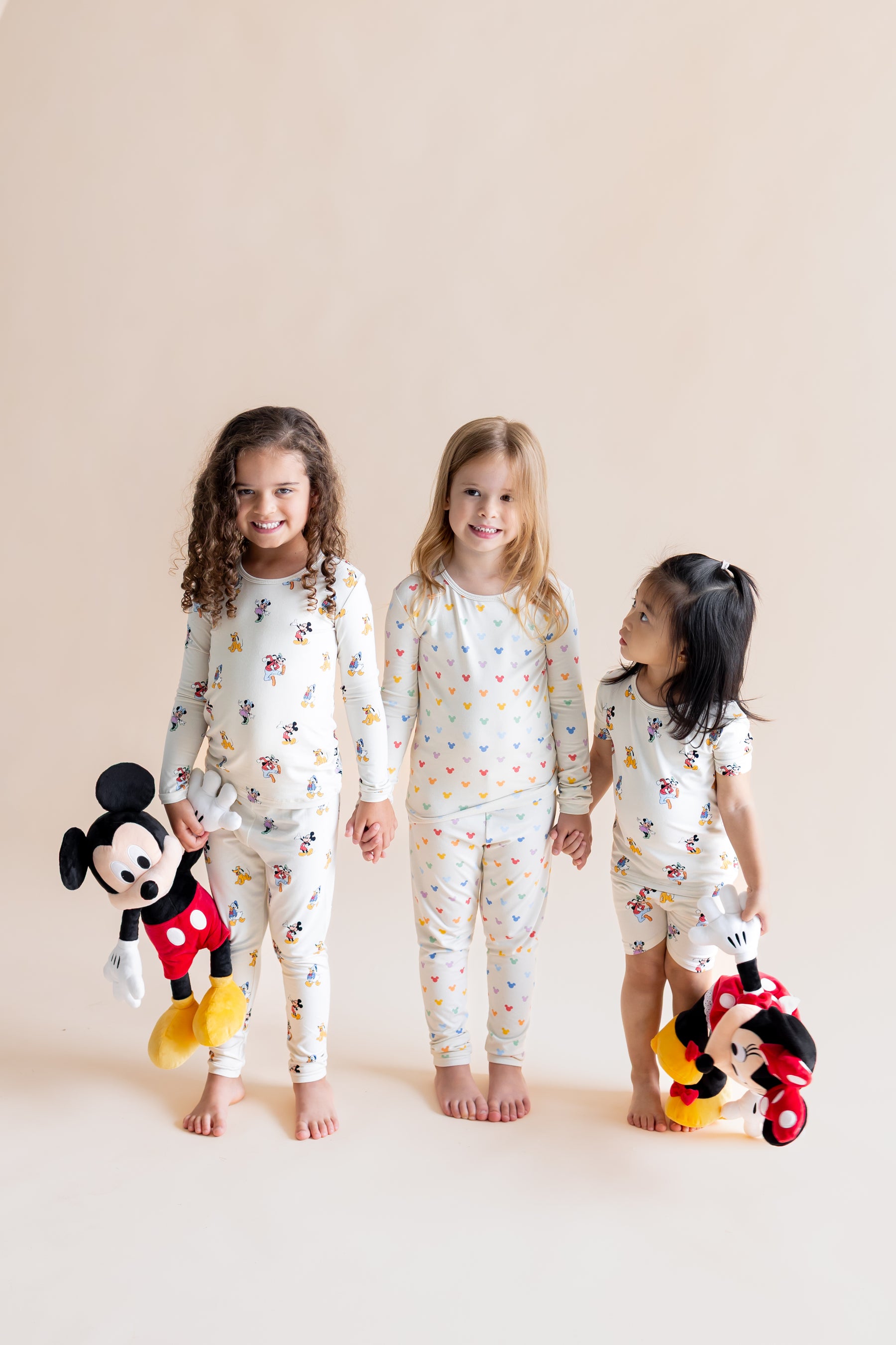 3 Girls standing holding each others hands wearing Long Sleeve Pajamas in in Mickey Mouse & Friends and Pastel Mickey Mouse print and Short Sleeve Pajamas in Mickey Mouse & Friends print holding stuff toys on a white background