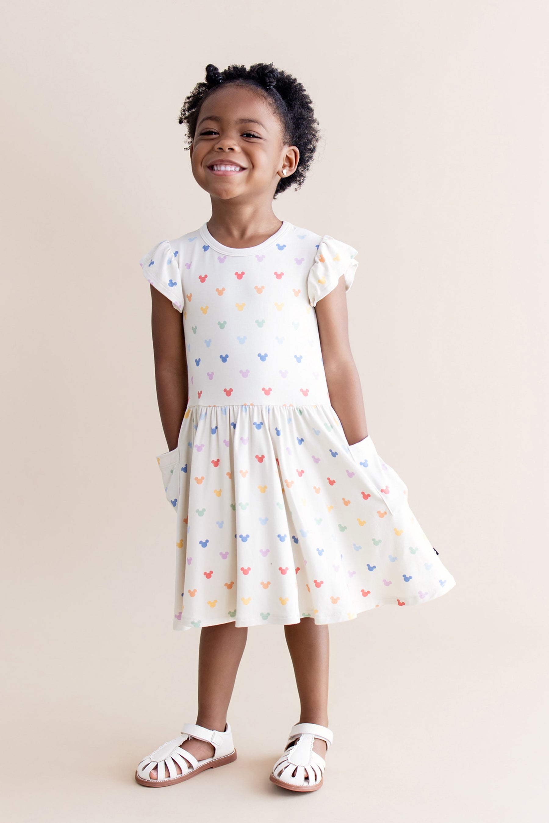 Young girl wearing a cream pocket dress with colorful mickey patterns on a beige background