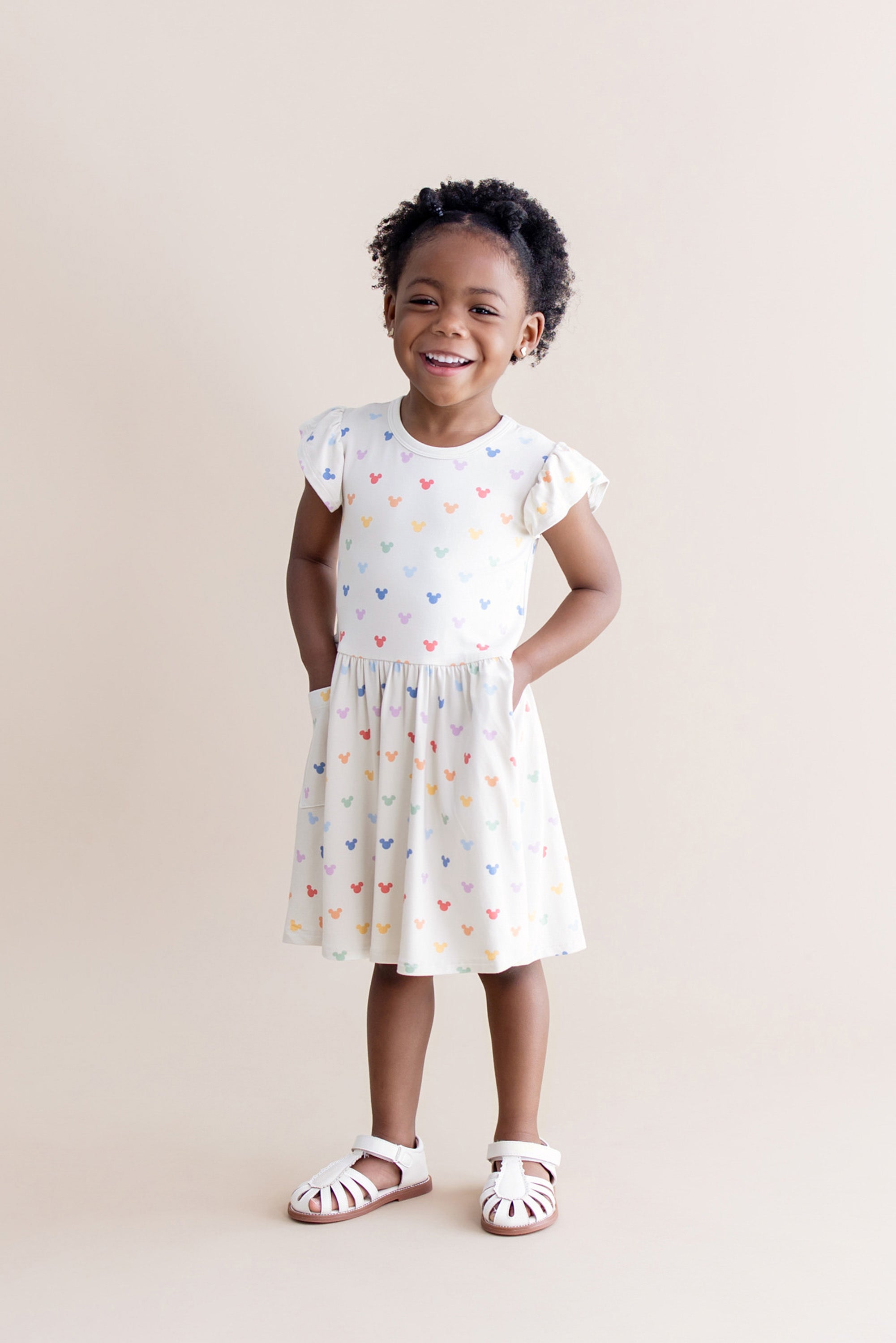 Young girl wearing a cream dress with colorful mickey mouse head patterns on a beige background