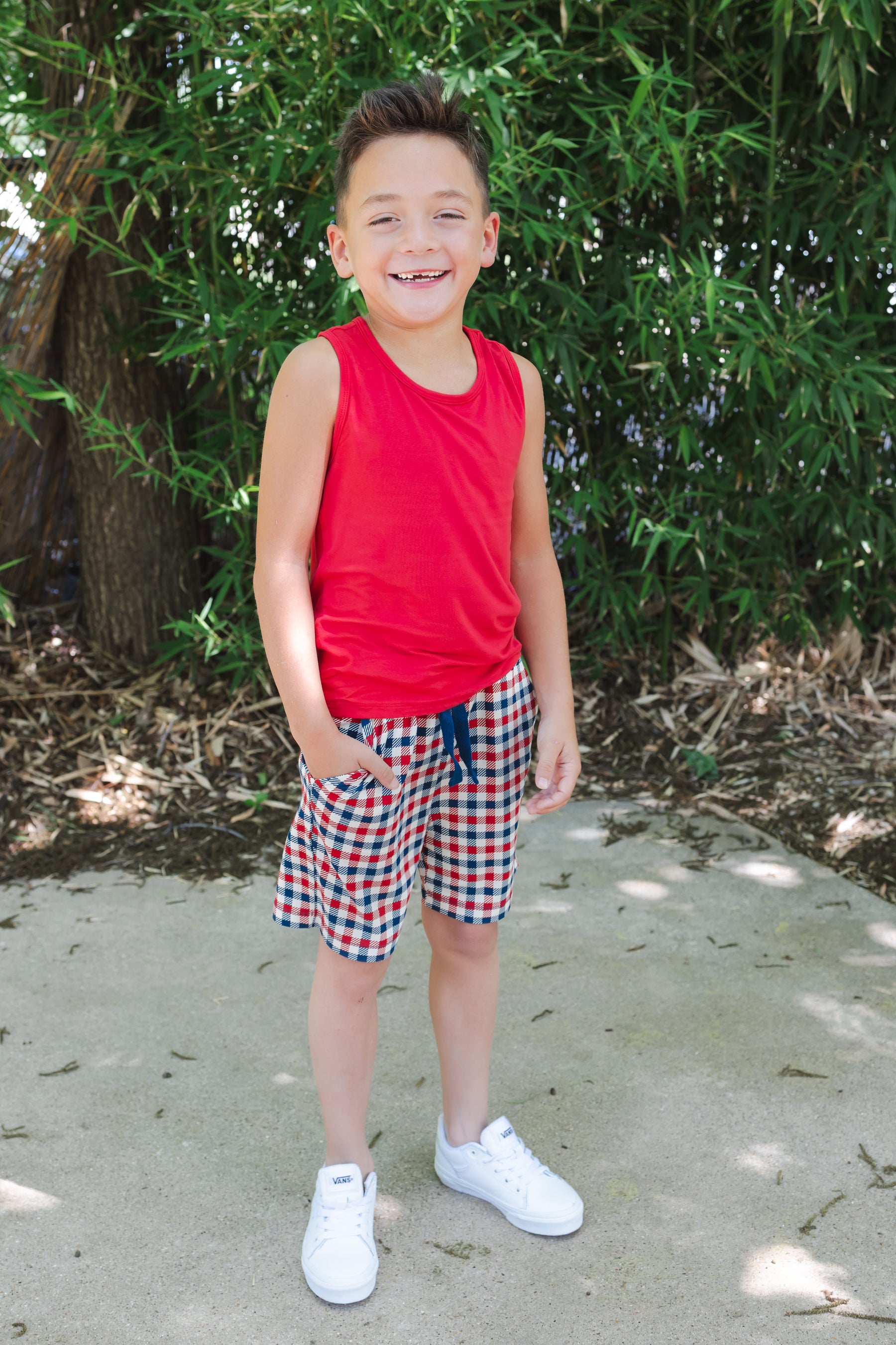 Child wearing a red blue and white Patriotic Gingham check print drawstring shorts outdoors over a red tank top and white shoes