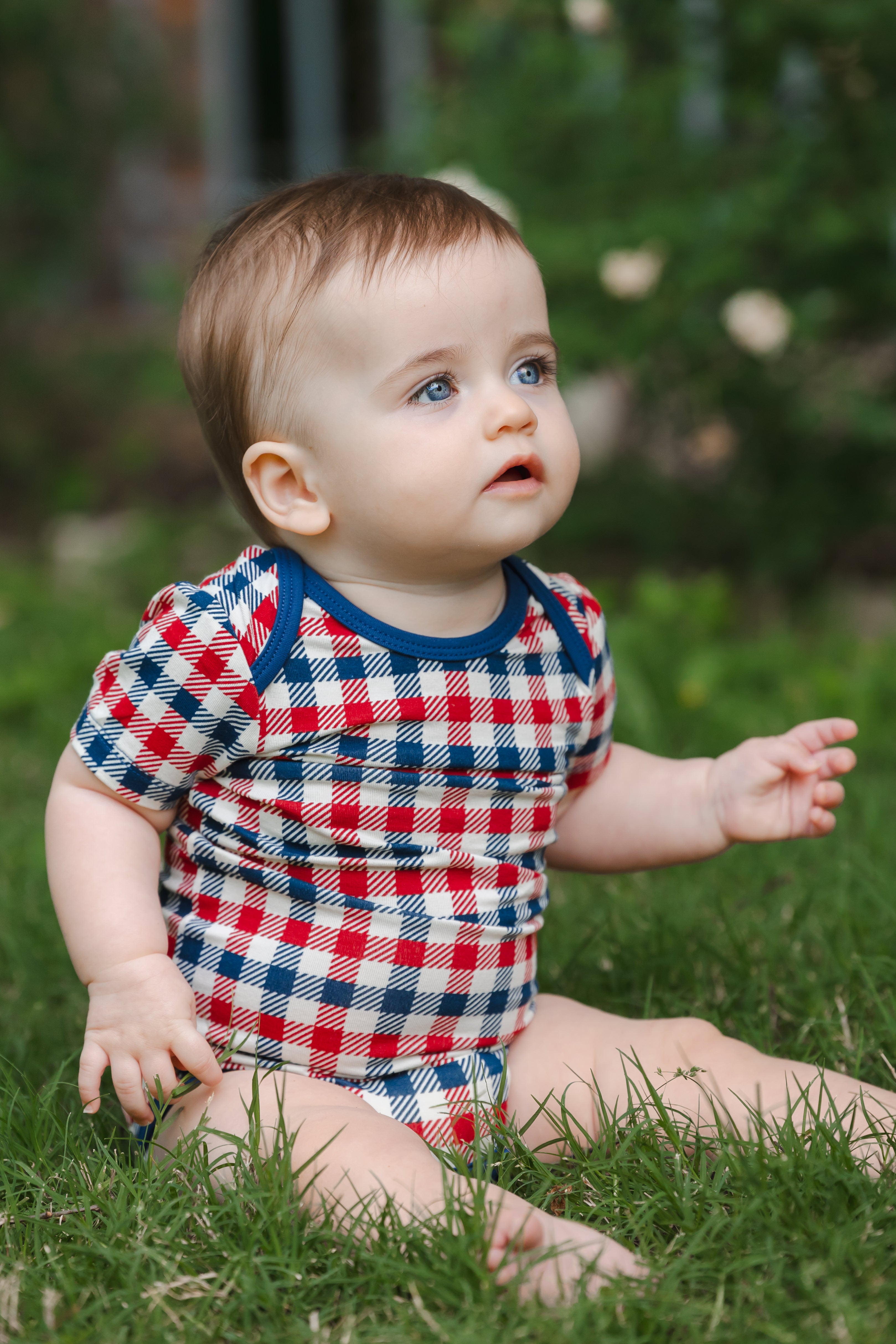 Baby wearing a short sleeve Bodysuit in red blue and white Patriotic Gingham check print sitting on a grass