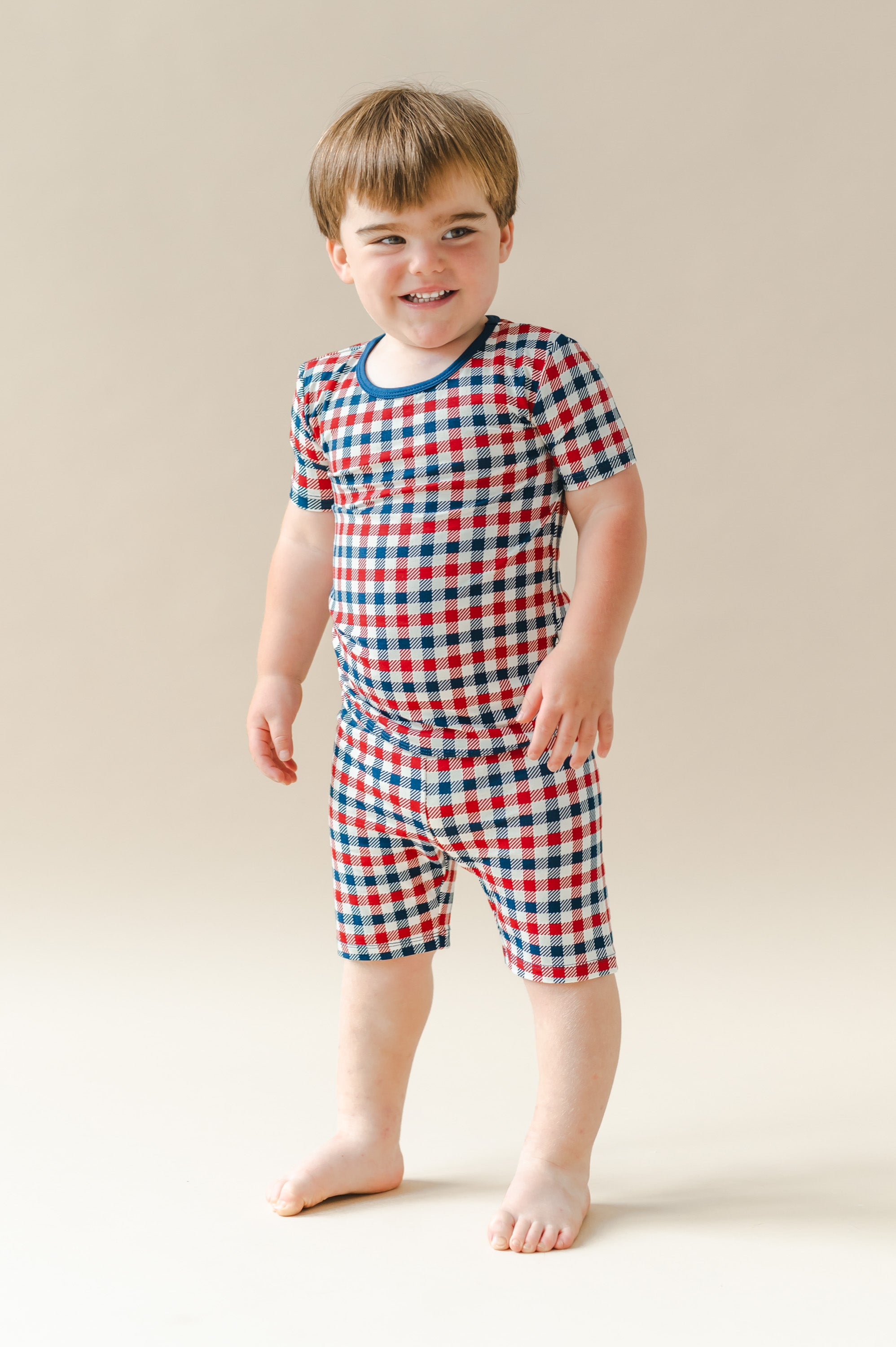 Child standing in Short Sleeve Pajamas in red blue and white Patriotic Gingham check print on a beige background