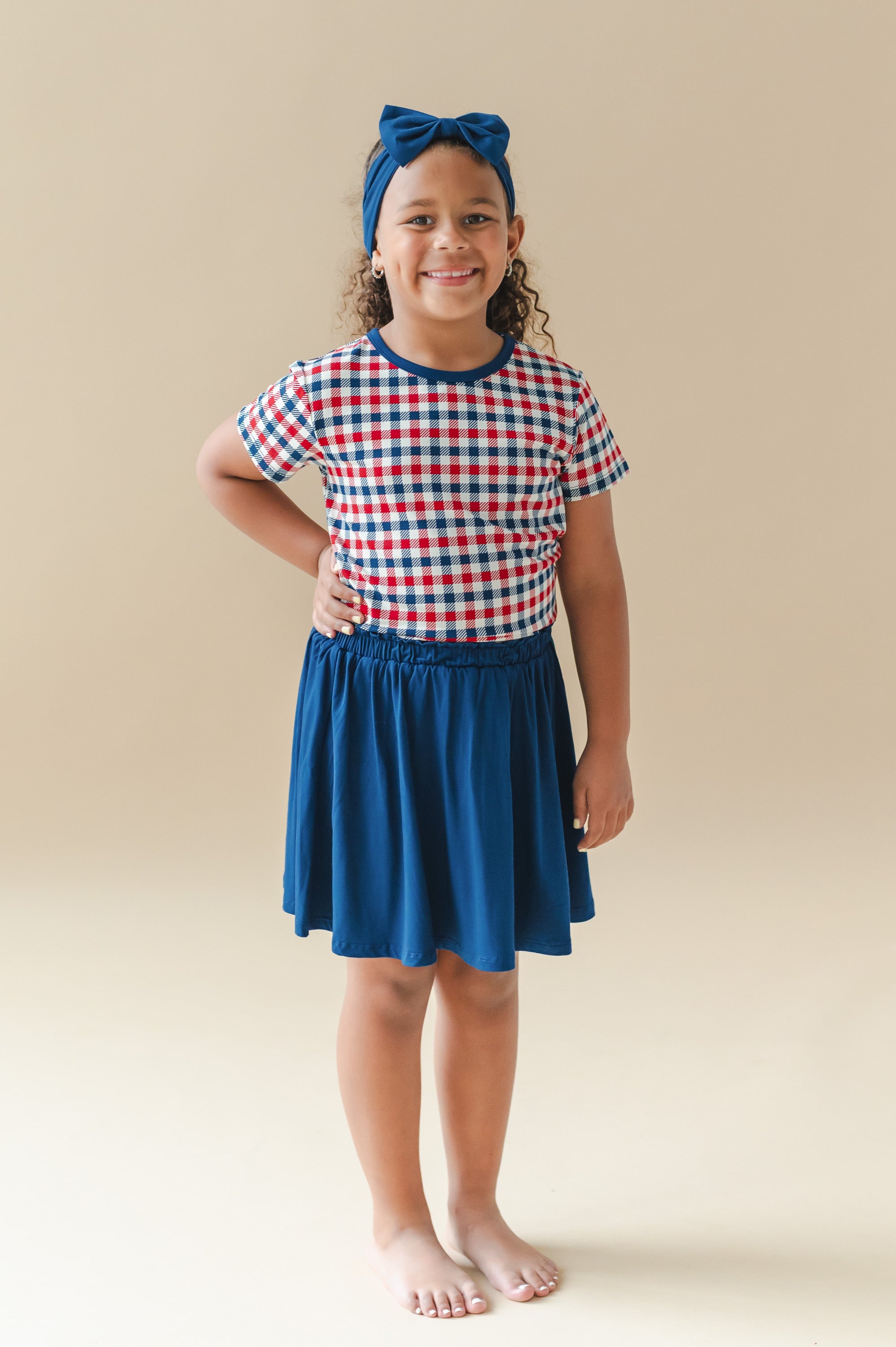 Child wearing Toddler Basic Tee in red blue and white Patriotic Gingham check print with a blue skort and bow headband on a beige background