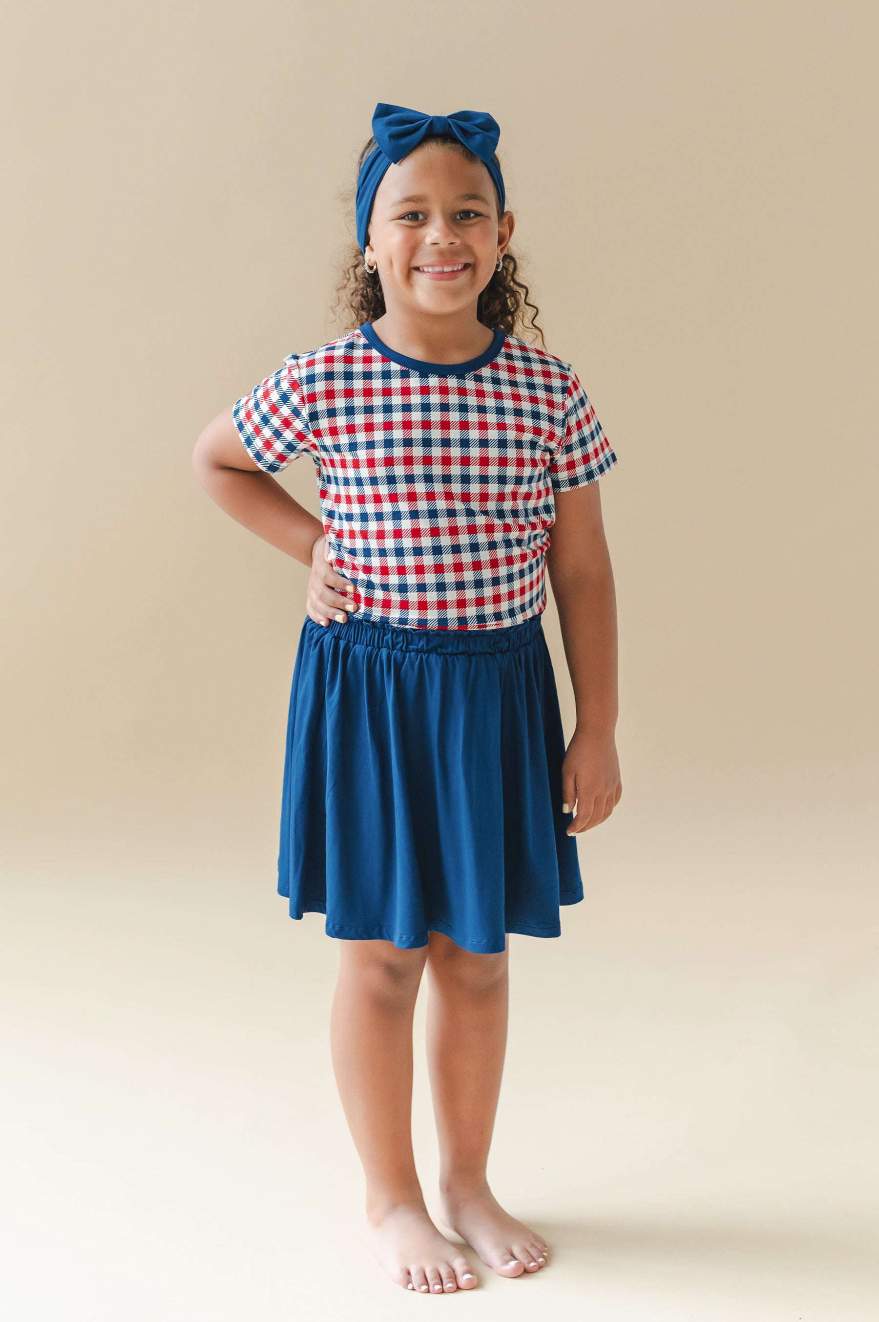 Child wearing Toddler Basic Tee in red blue and white Patriotic Gingham check print with a blue skort and bow headband on a beige background