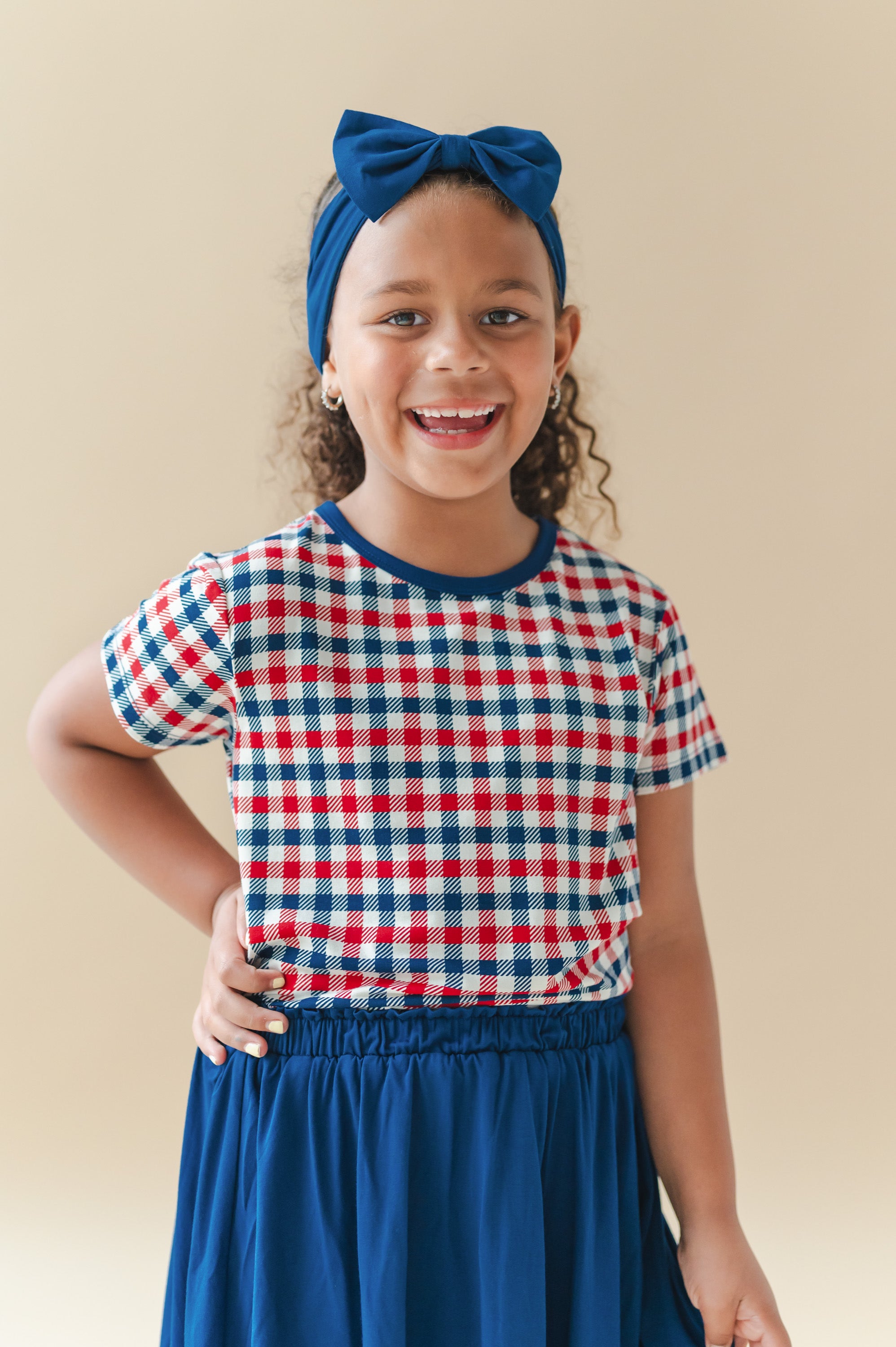 Child wearing Toddler Basic Tee in red blue and white Patriotic Gingham check print over a Tahoe skort and bow headband on a beige background