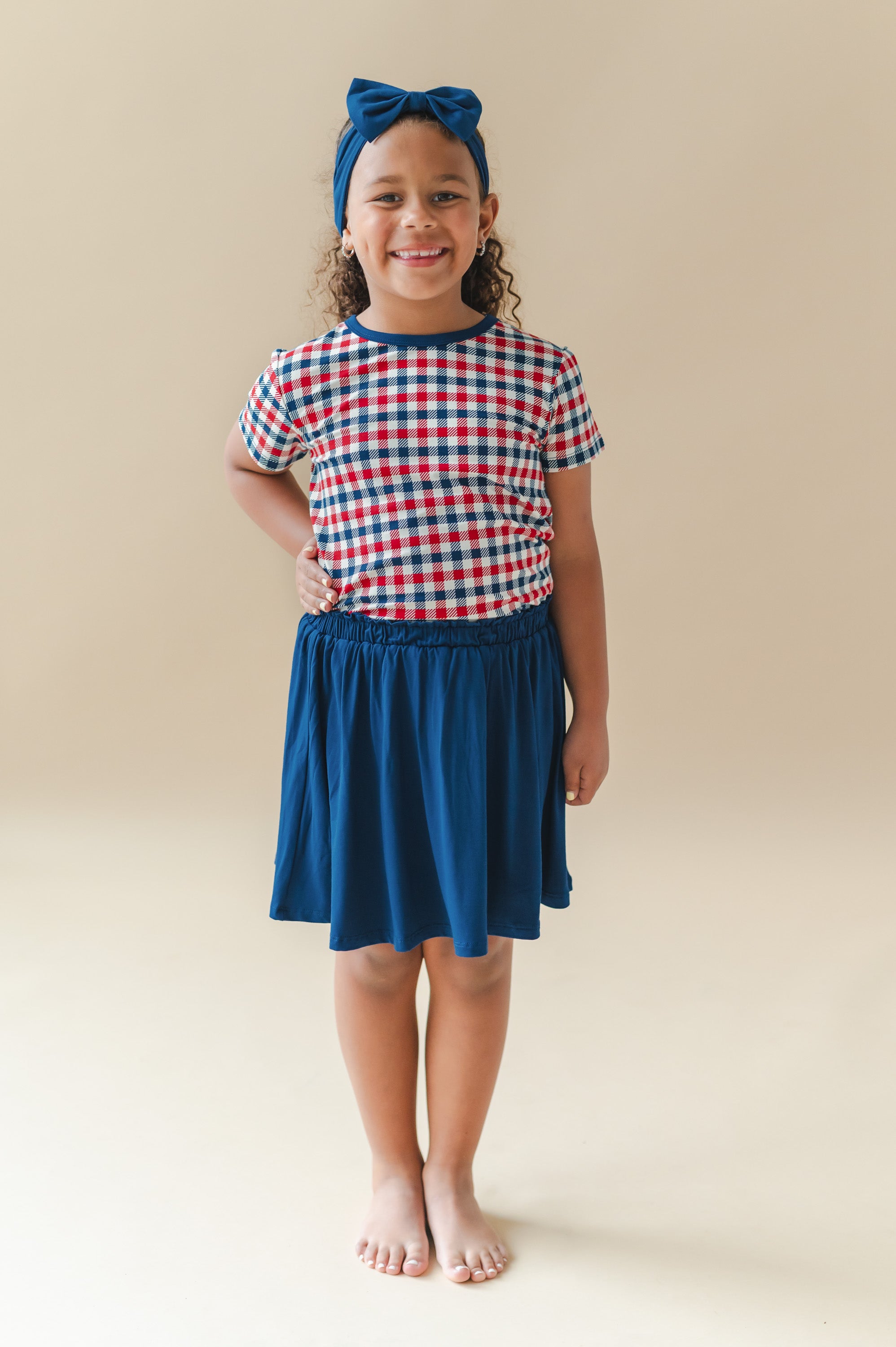 Child wearing Toddler Basic Tee in red blue and white Patriotic Gingham check print with a Tahoe skort and bow headband on a beige background