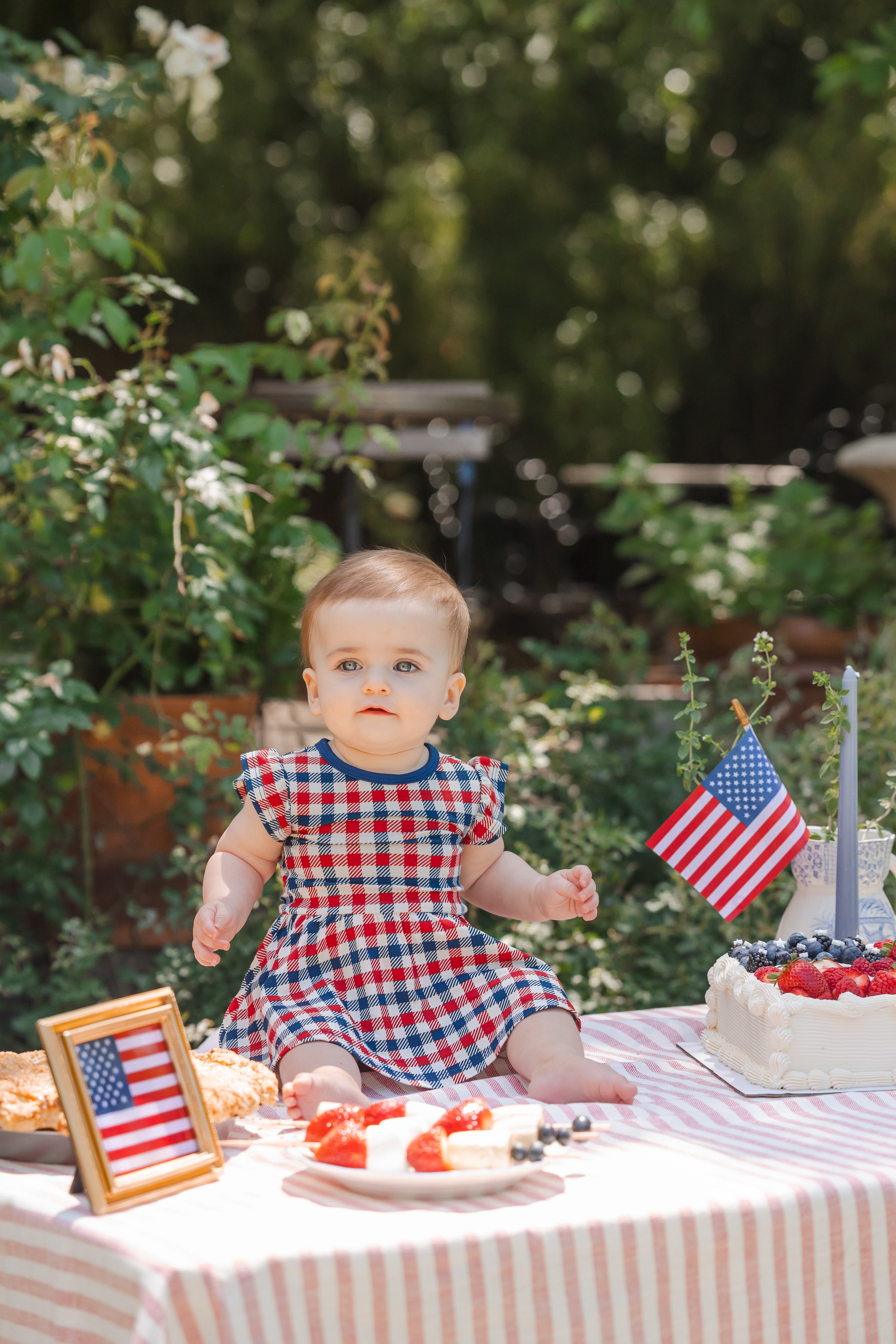 Child sitting outdoors on a table wearing a red blue and white Patriotic Gingham check print Twirl bodysuit dress