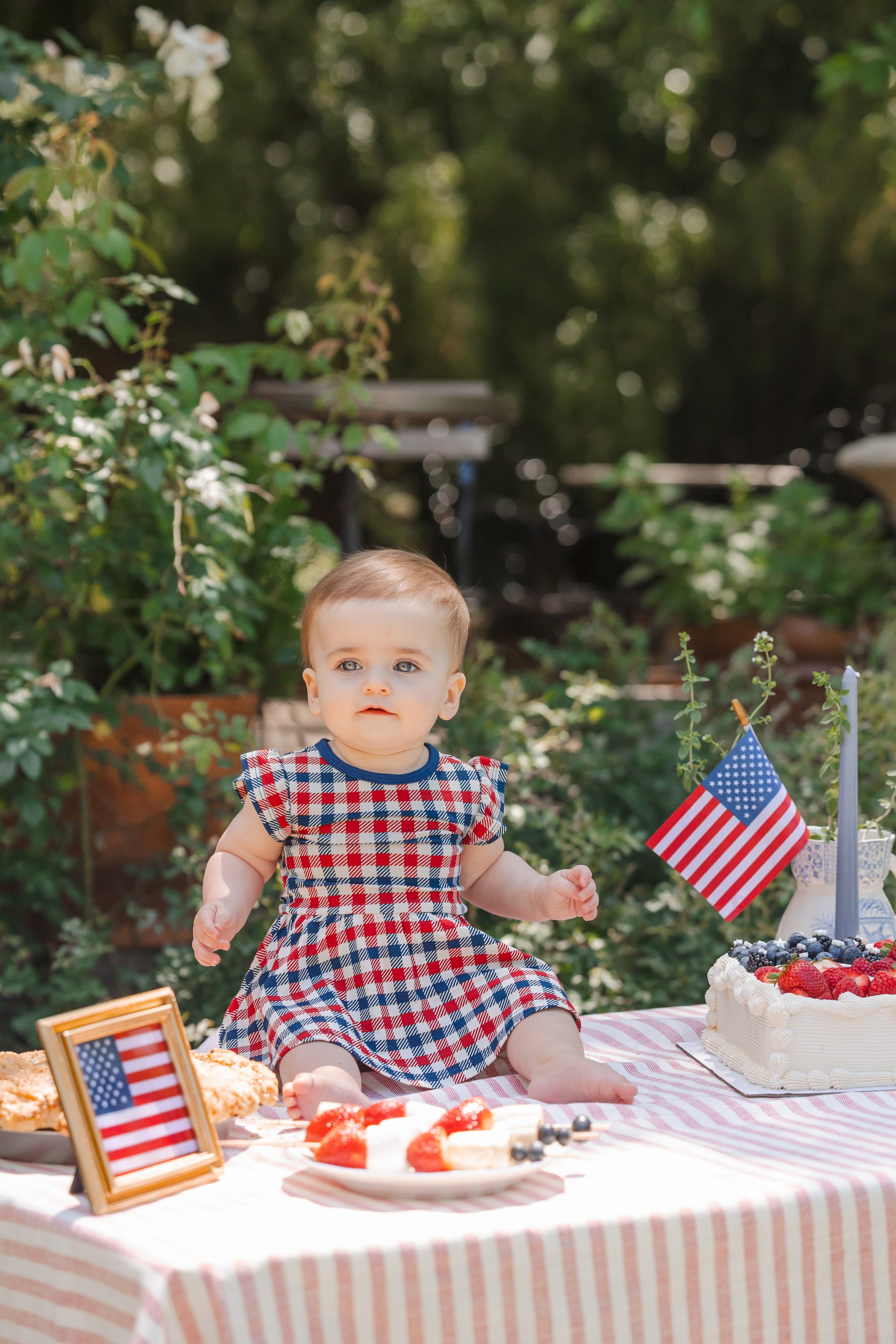 Child sitting outdoors on a table wearing a red blue and white Patriotic Gingham check print Twirl bodysuit dress