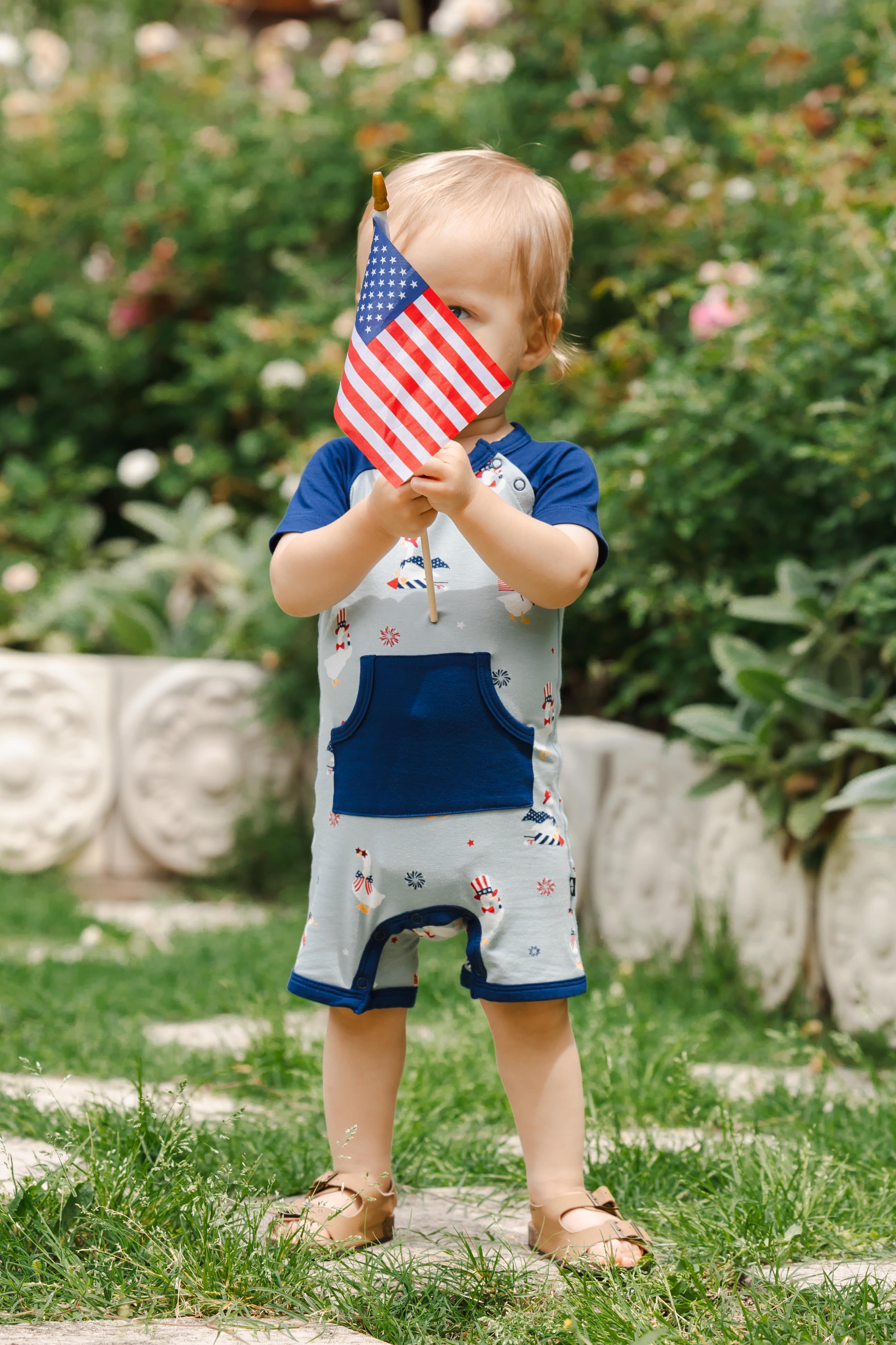 Child wearing light blue Shortall in Patriotic Goose print with navy trim holding a flag outdoors
