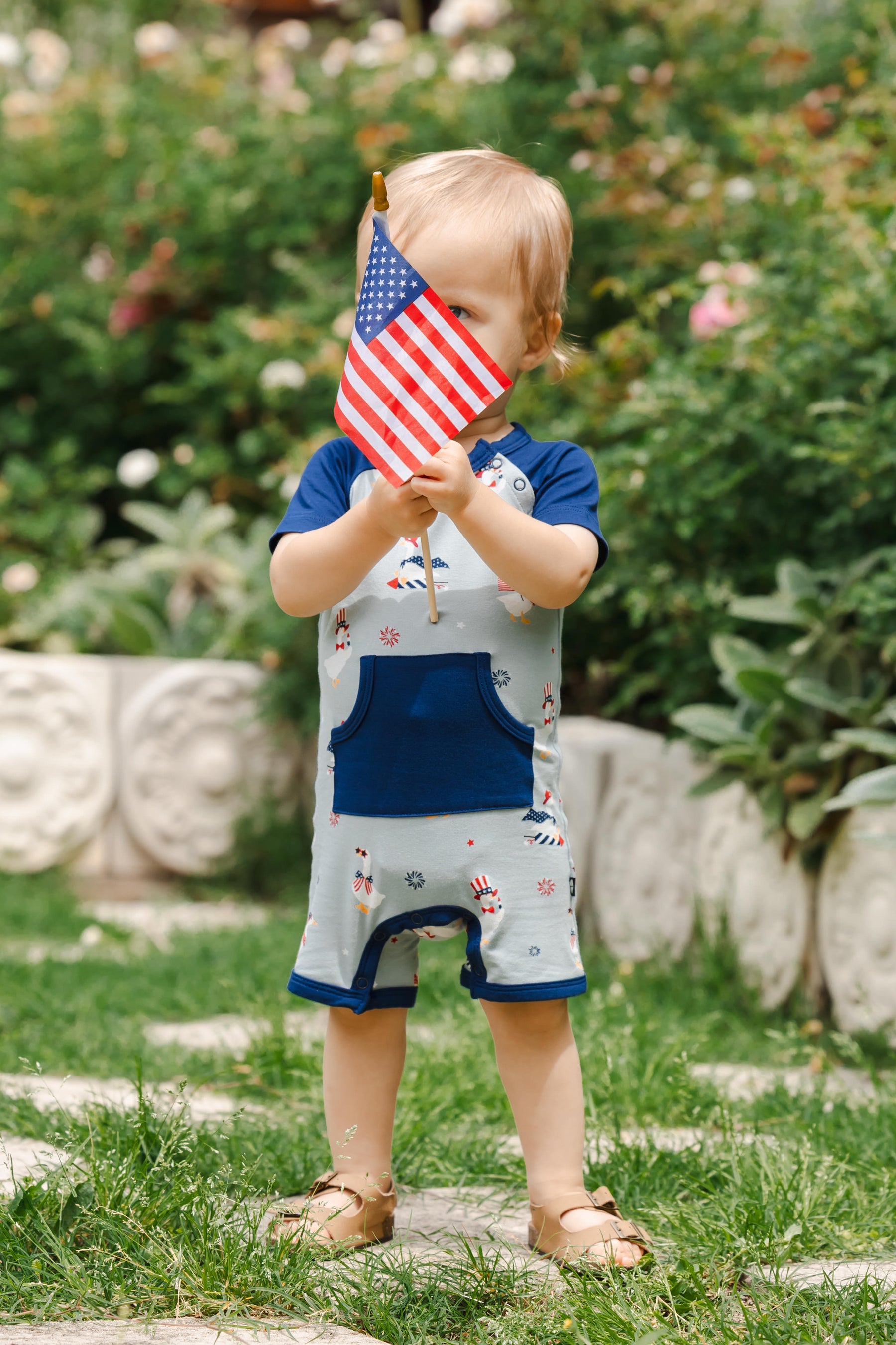 Child wearing light blue Shortall in Patriotic Goose print with navy trim holding a flag outdoors