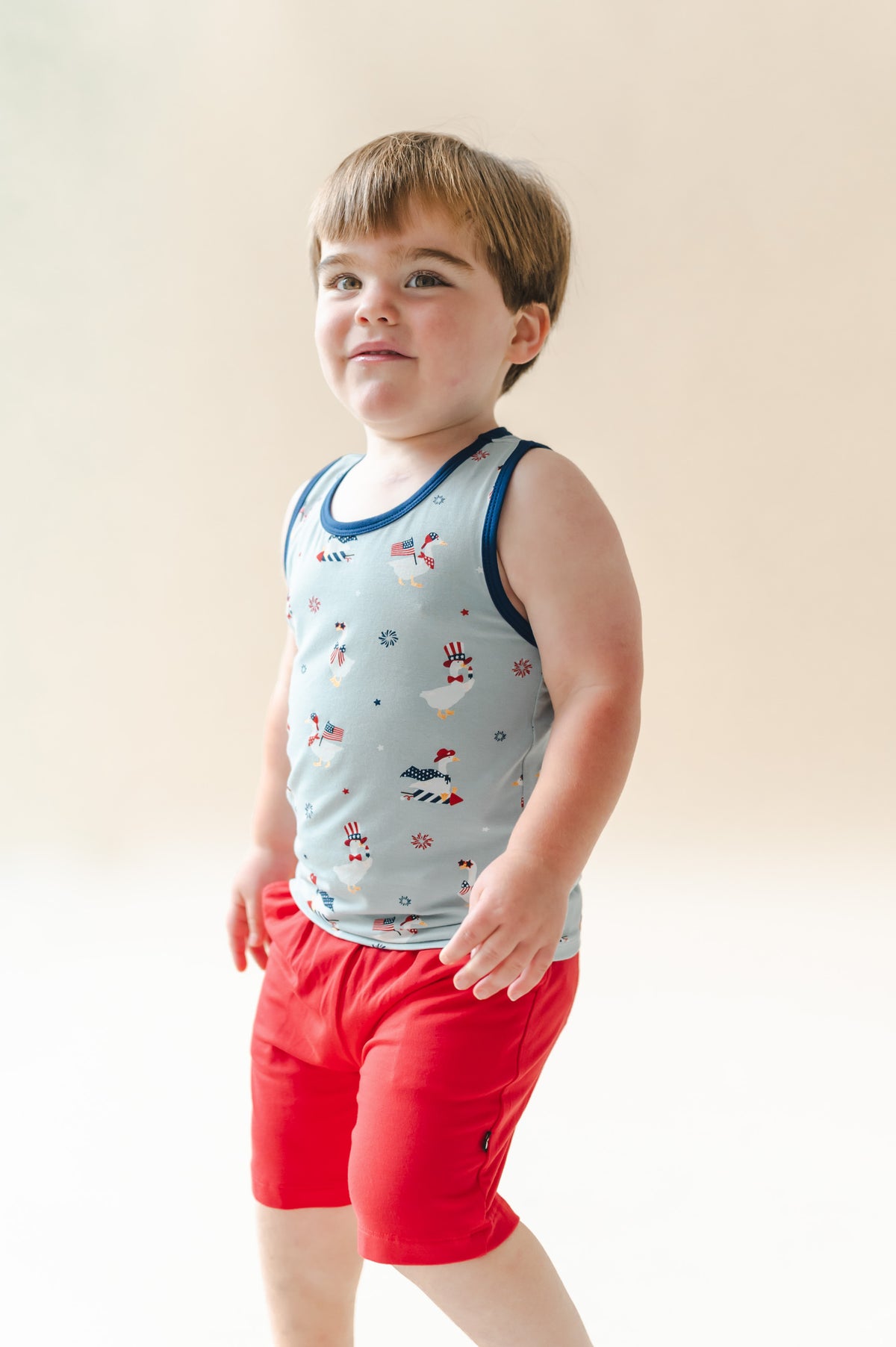 Child wearing a light blue Toddler tank top in Patriotic Goose print with navy trim over red shorts on a beige background