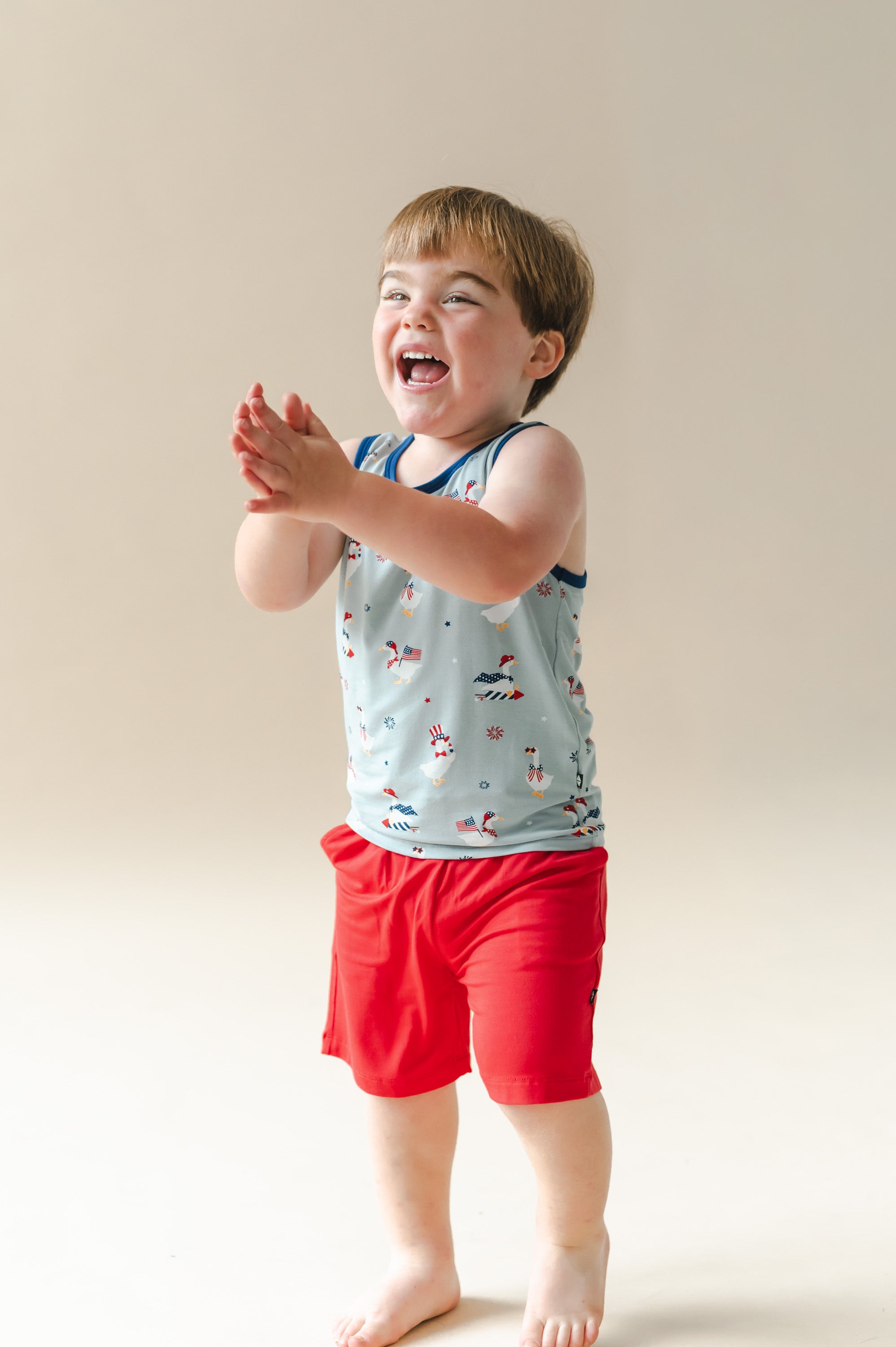 Child laughing in light blue Toddler tank top in Patriotic Goose print with navy trim over red shorts on a beige background