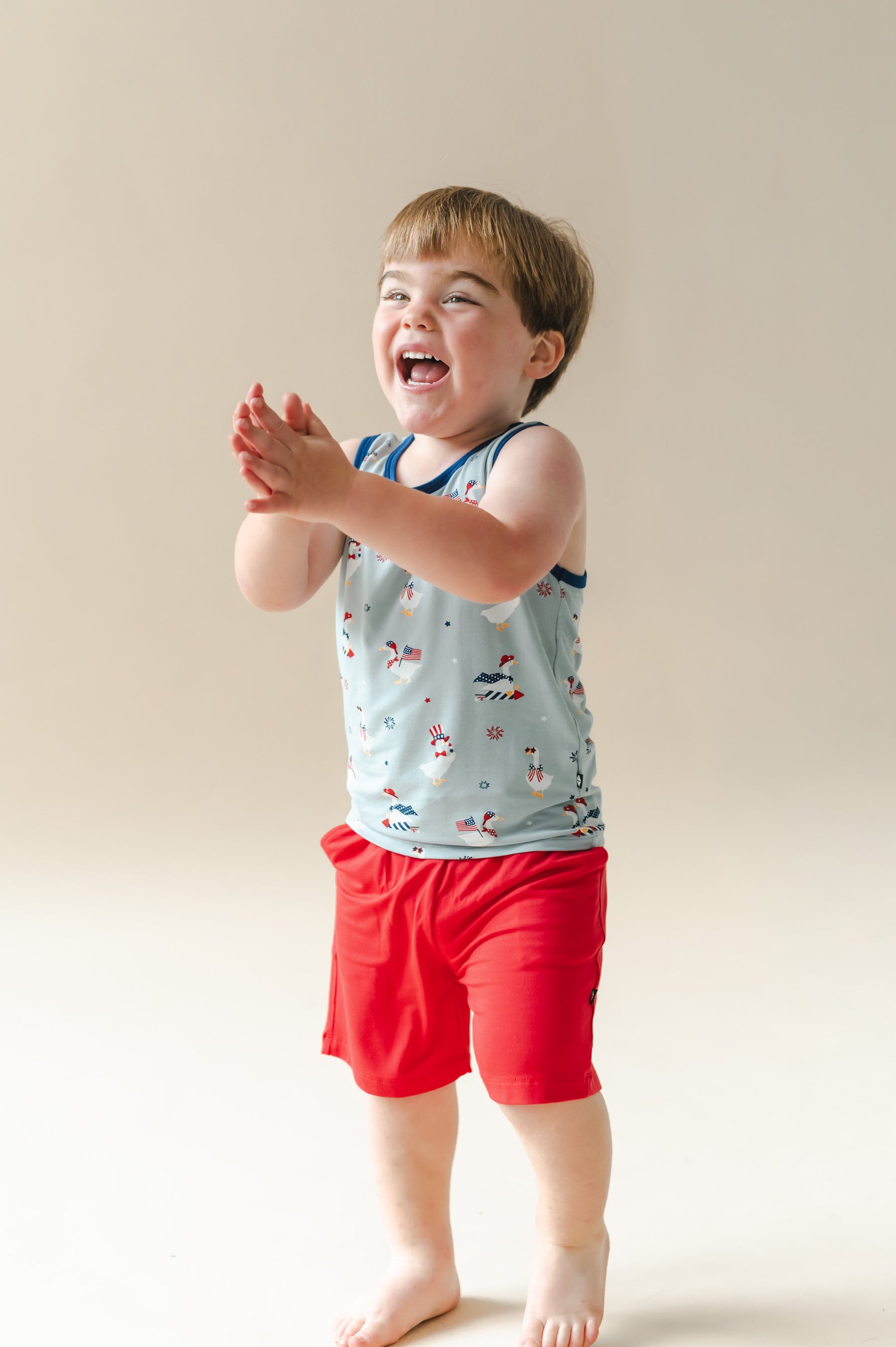 Child laughing in light blue Toddler tank top in Patriotic Goose print with navy trim over red shorts on a beige background