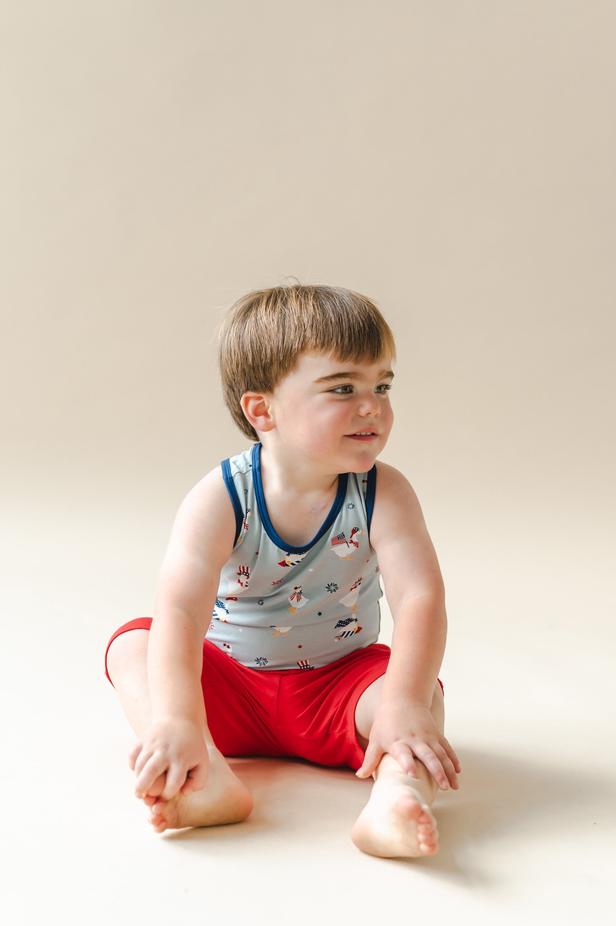 Child sitting in a Light blue Toddler tank top in Patriotic Goose print with navy trim over red shorts on a beige background
