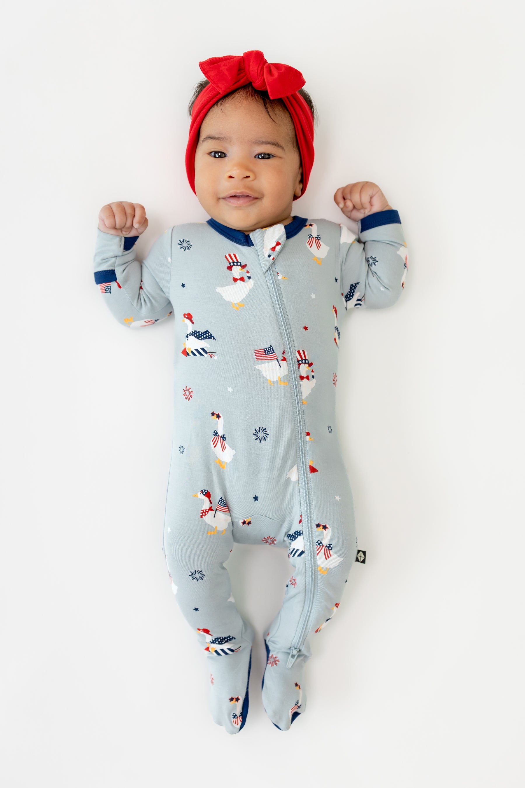 Baby wearing Light blue Zippered footie in Patriotic Goose print with a red bow headband on a white background