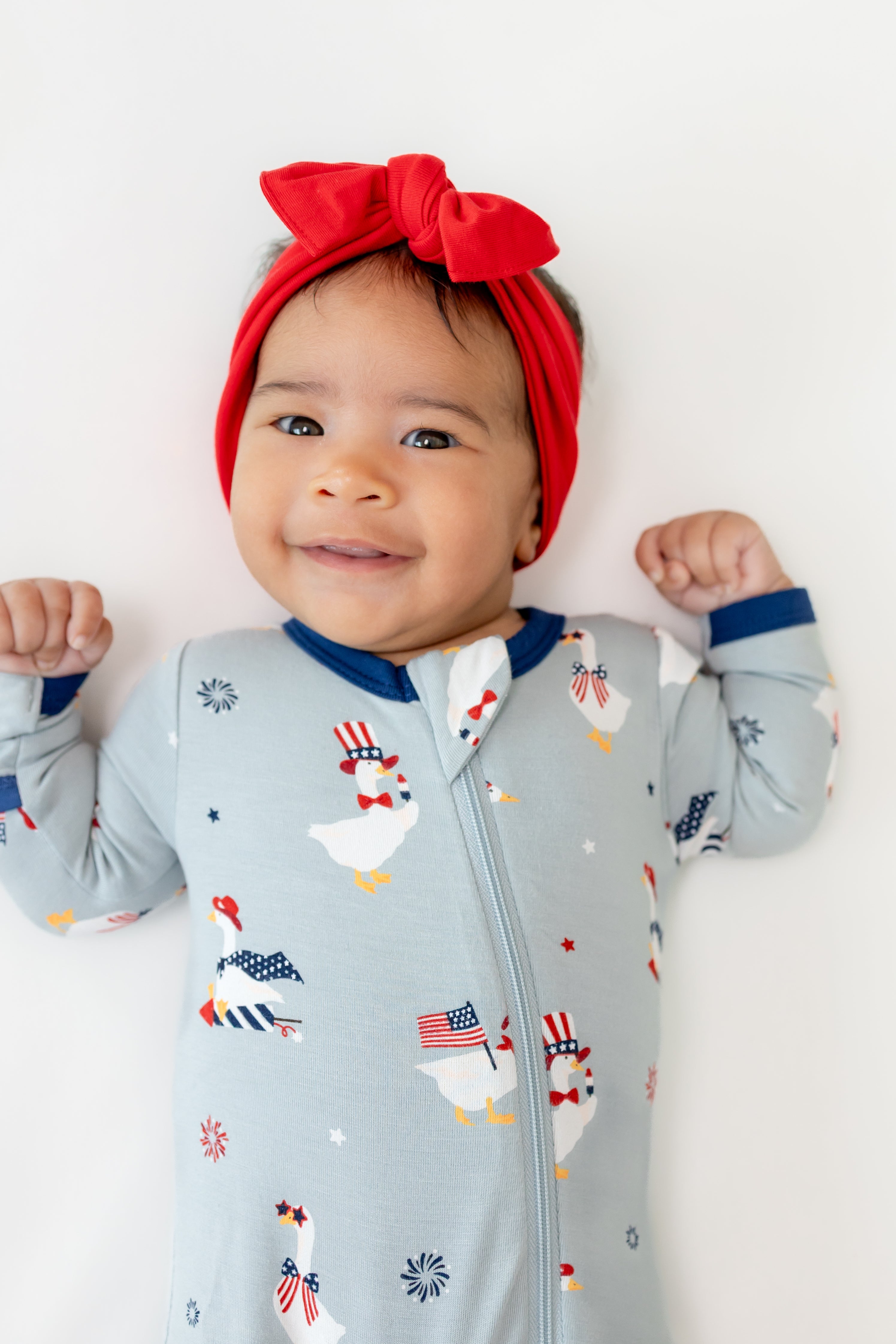 Baby wearing Light blue Zippered footie in Patriotic Goose print with a red bow headband on a white background