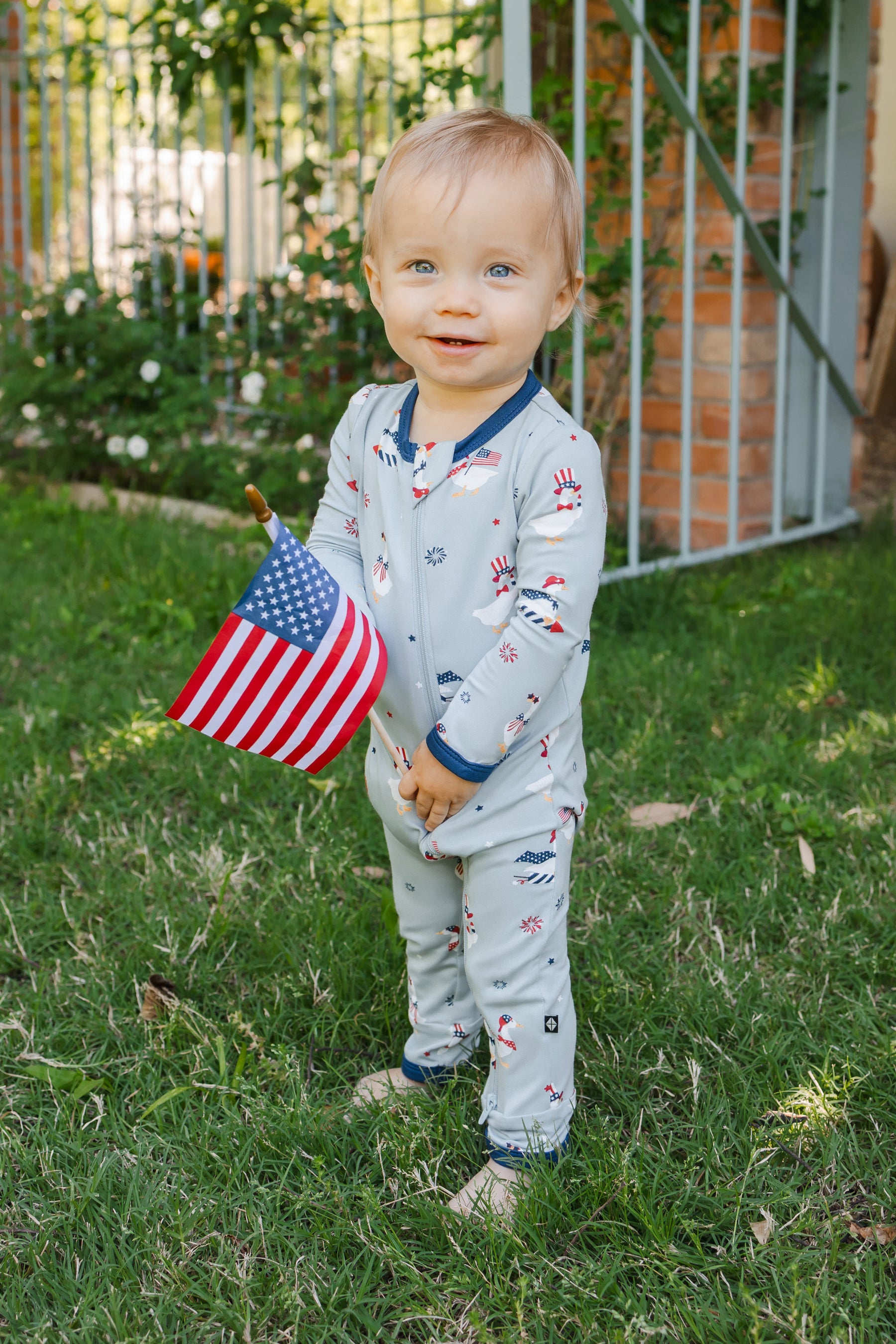 Child standing wearing a light blue zippered romper in Patriotic Goose print with a flag outdoors