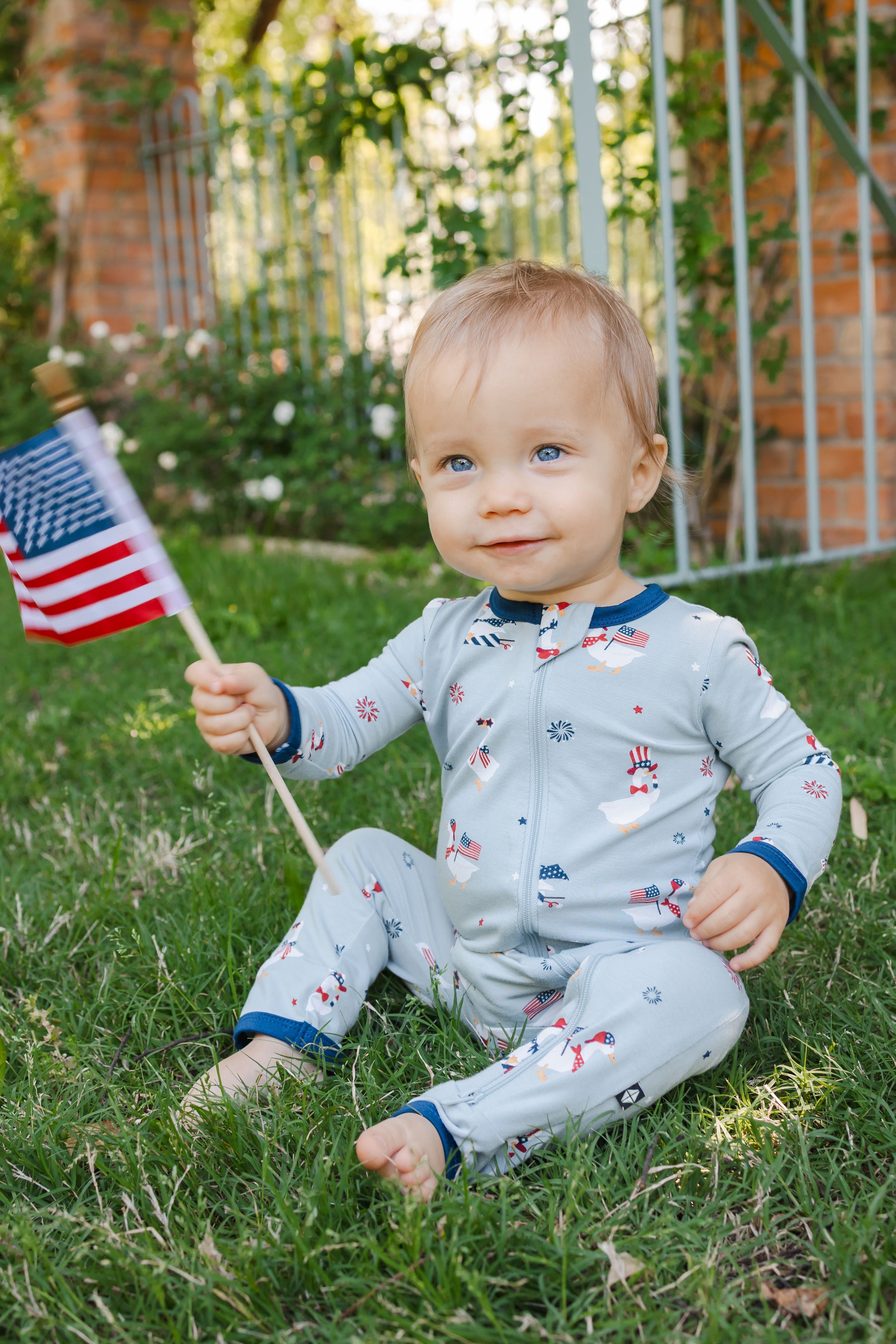 Child sitting in Patriotic Goose print light blue zippered romper with a flag outdoors on a grass