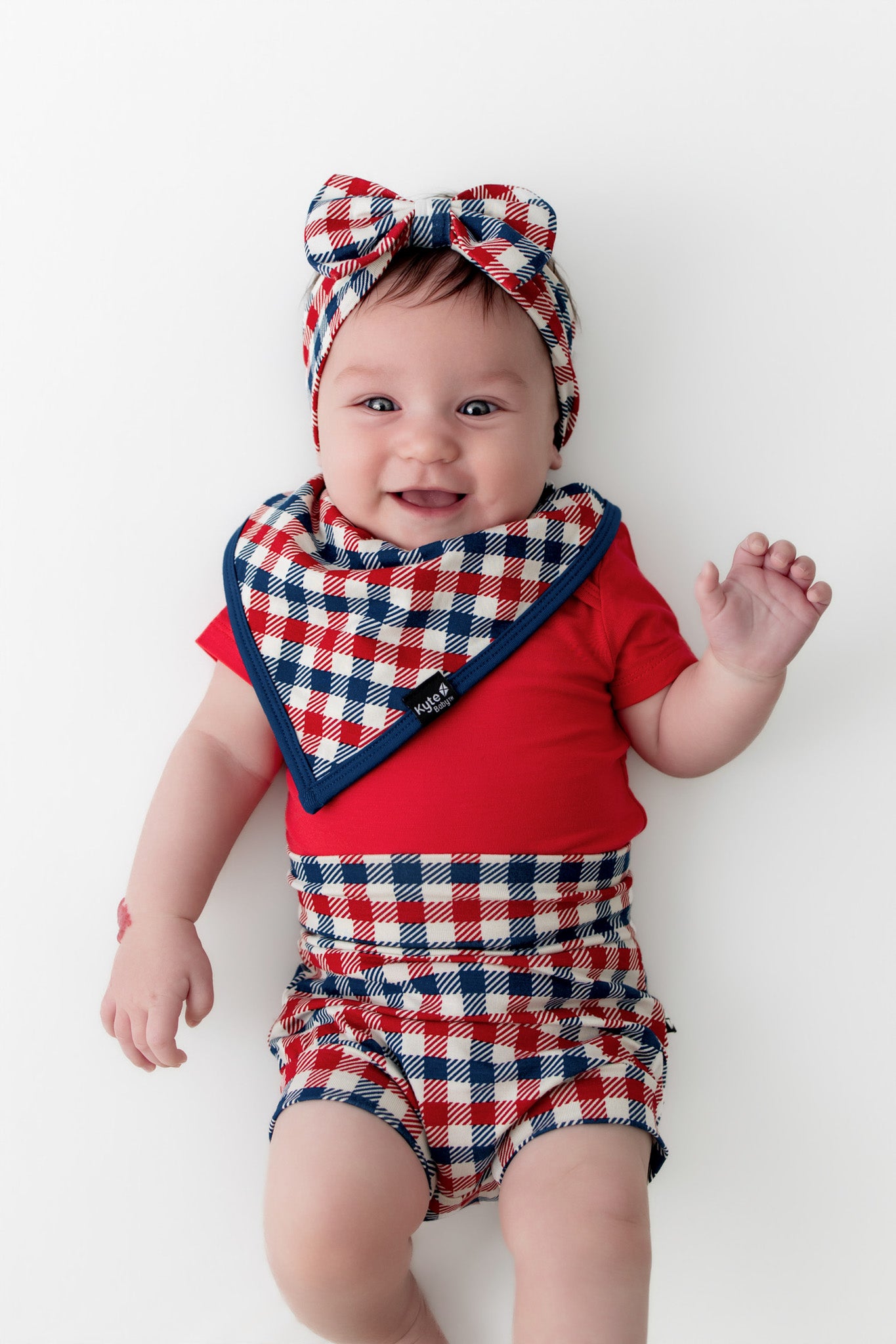 Baby wearing a red blue and white Patriotic Gingham check print bib with navy trim along with similar print bummies and bow headband over a red bodysuit on a white background