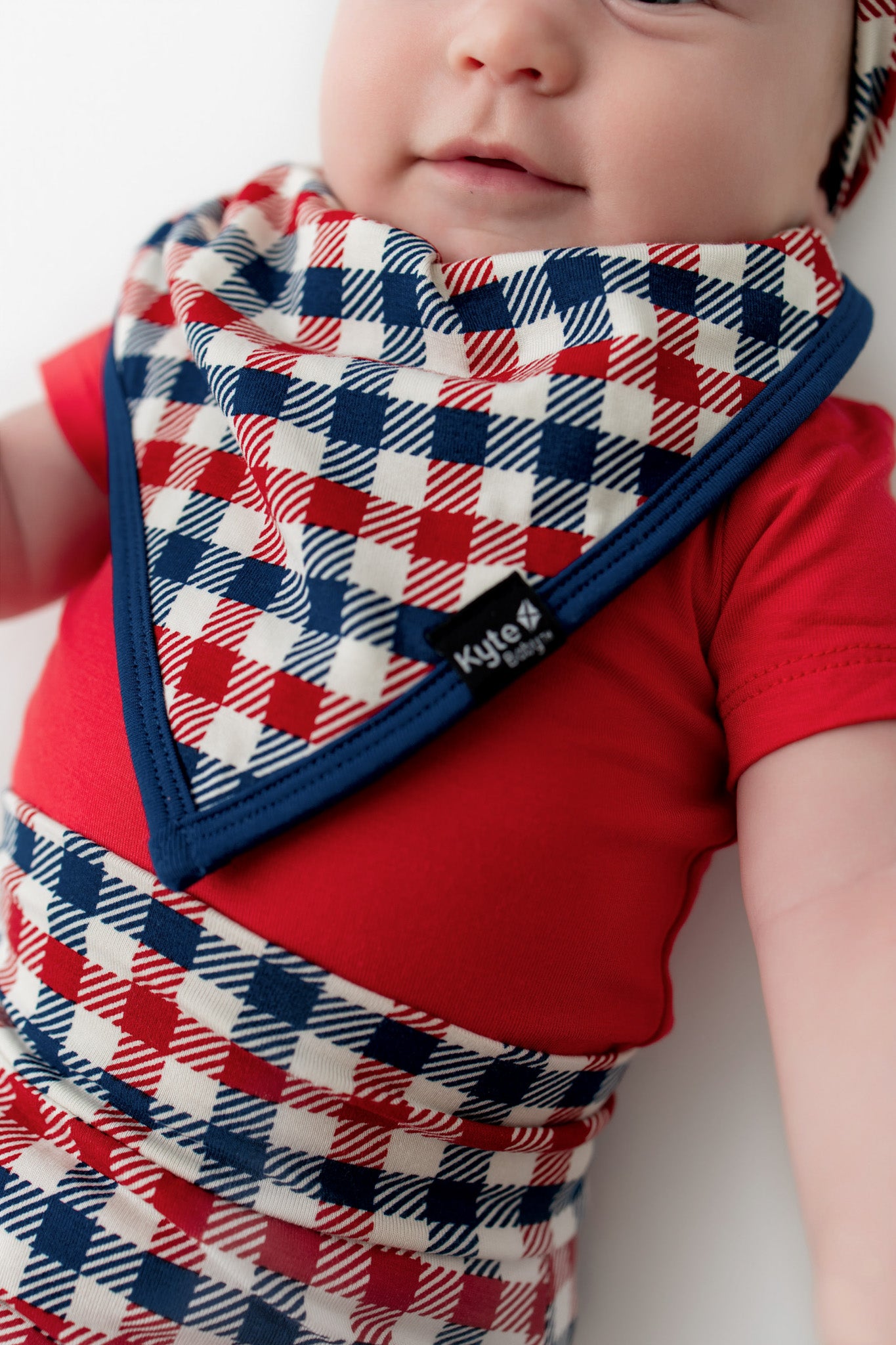 Baby wearing a red blue and white Patriotic Gingham check print bib with navy trim along with similar print bummies over a red bodysuit on a white background