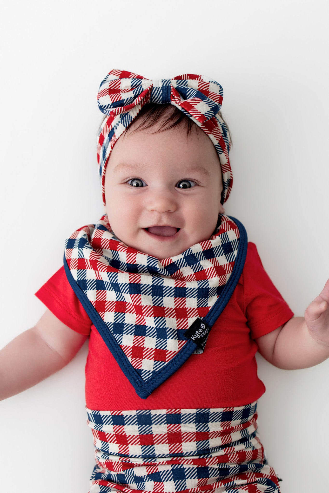 Baby wearing a bib in red blue and white Patriotic Gingham check print with a navy trim along with similar print bummies and bow headband over a red bodysuit on a white background