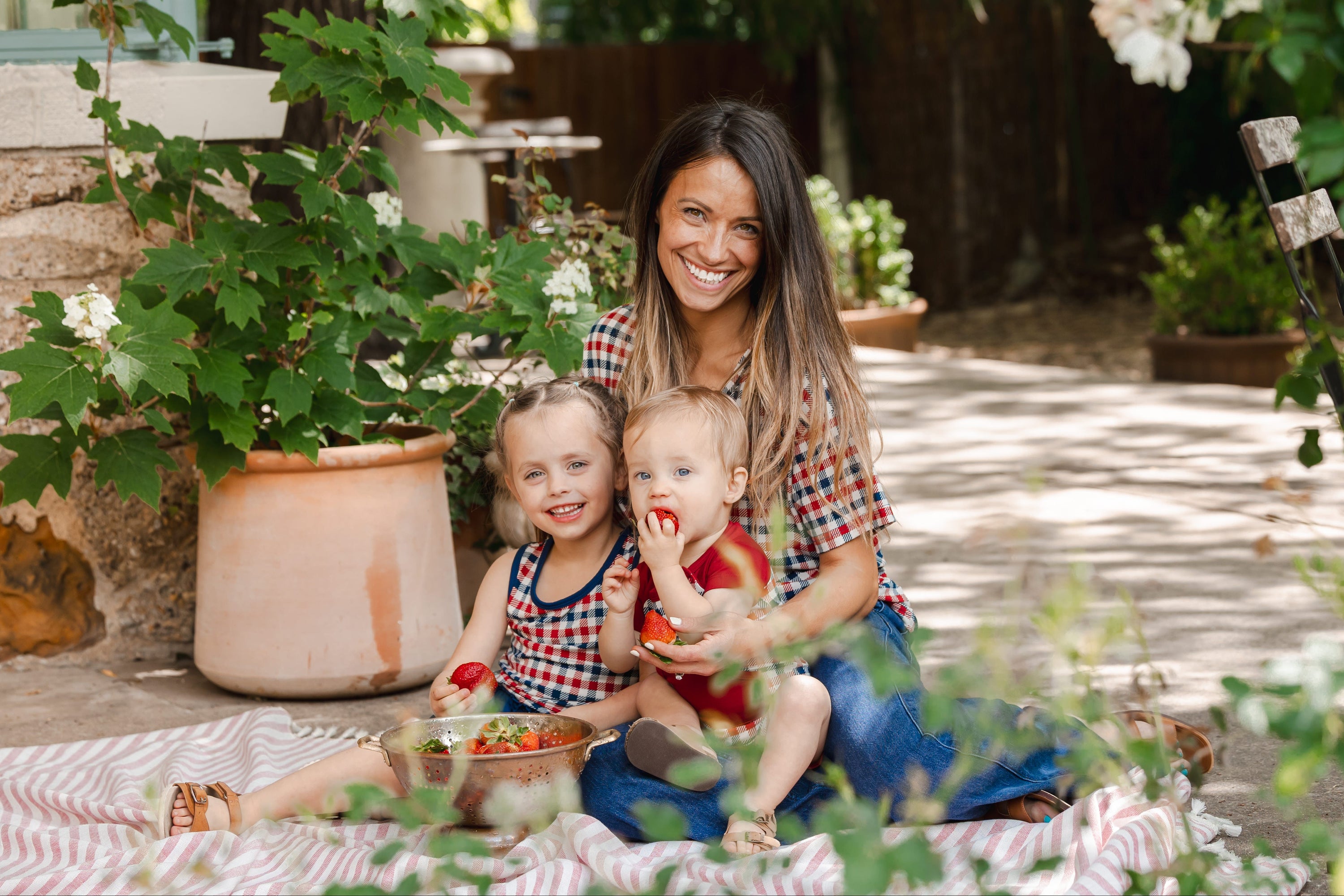 Woman and two children wearing matching red white and blue gingham apparel sitting on a blanket in a garden
