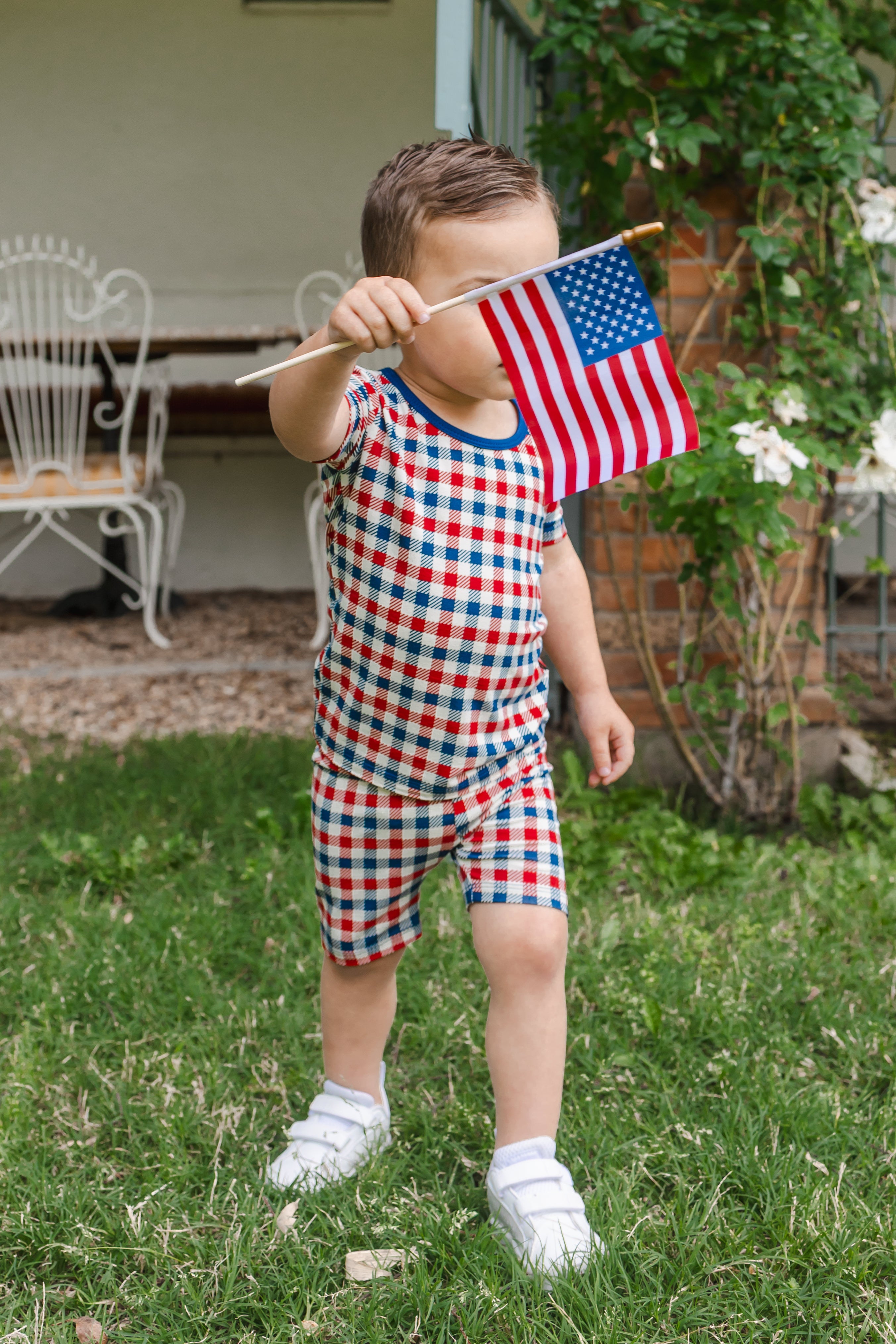 Child playing outdoors on a grass wearing a Short Sleeve Pajamas in red blue and white Patriotic Gingham check print with a flag
