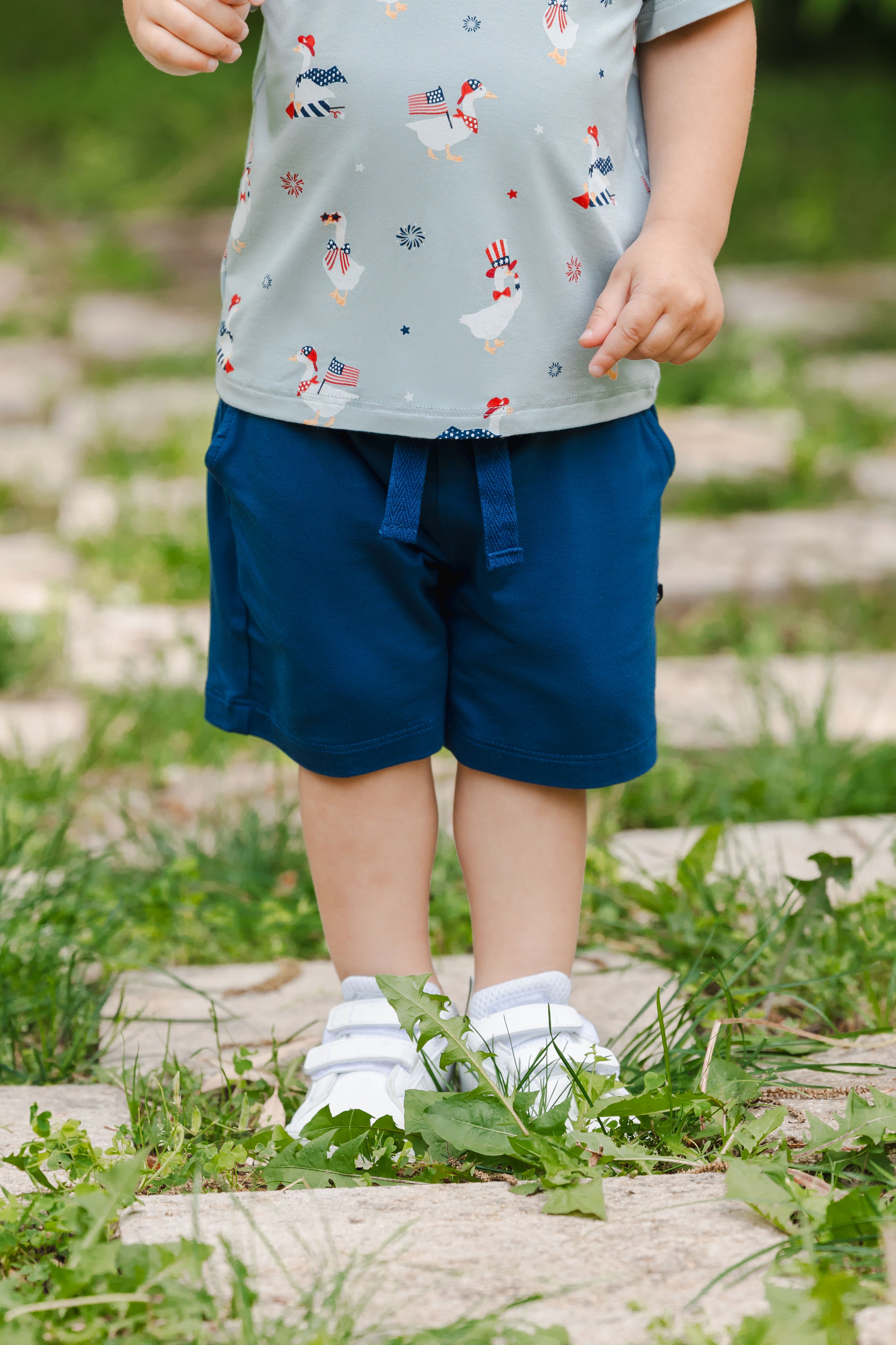 Toddler wearing Drawstring Short in Tahoe under blue shirt with patriotic goose print toddler v-neck tee and white shoes outdoors