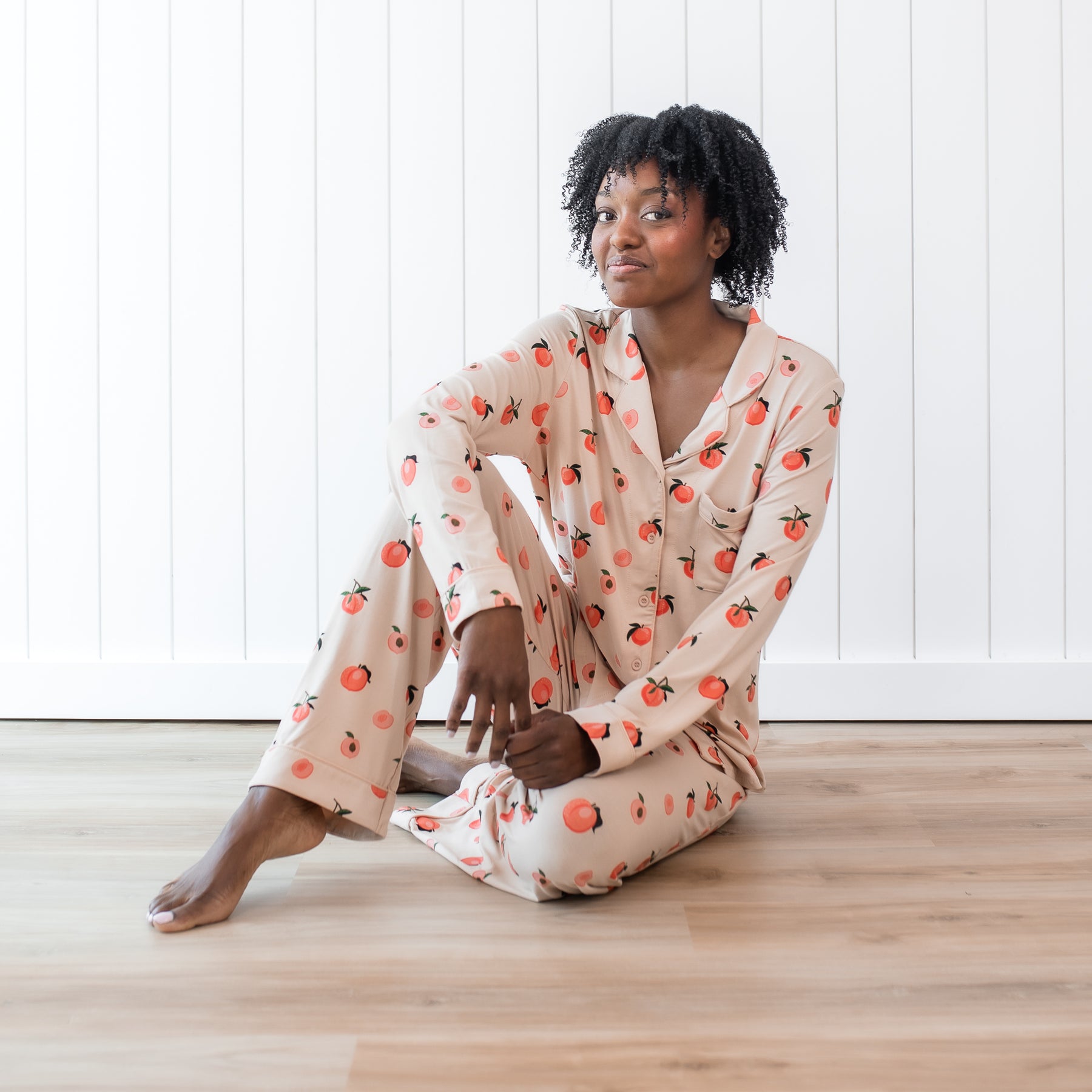 Woman modeling light pink button up bamboo pajamas in a peach print against white wooden wall