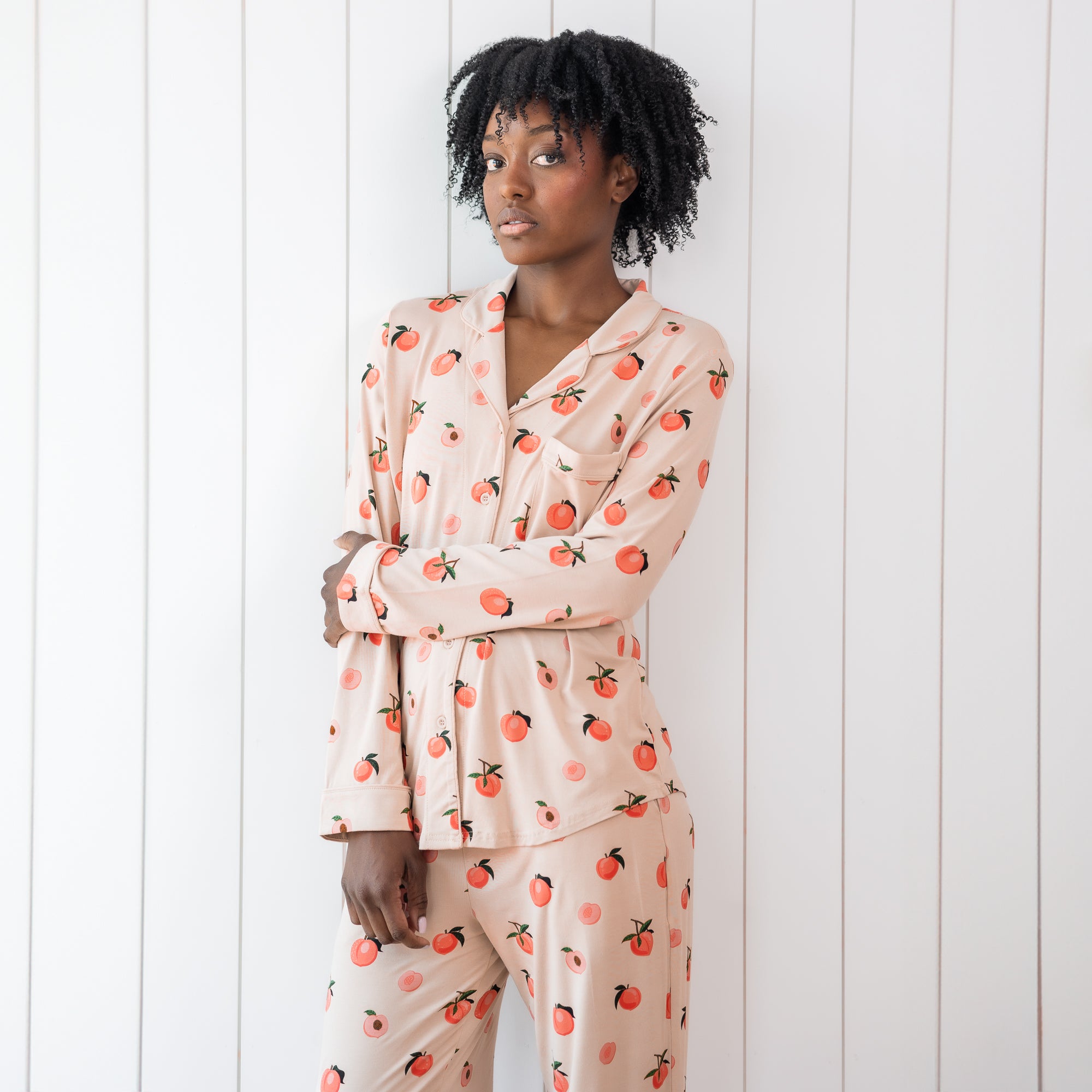 Woman modeling light pink button up bamboo pajamas in a peach print against white wooden wall